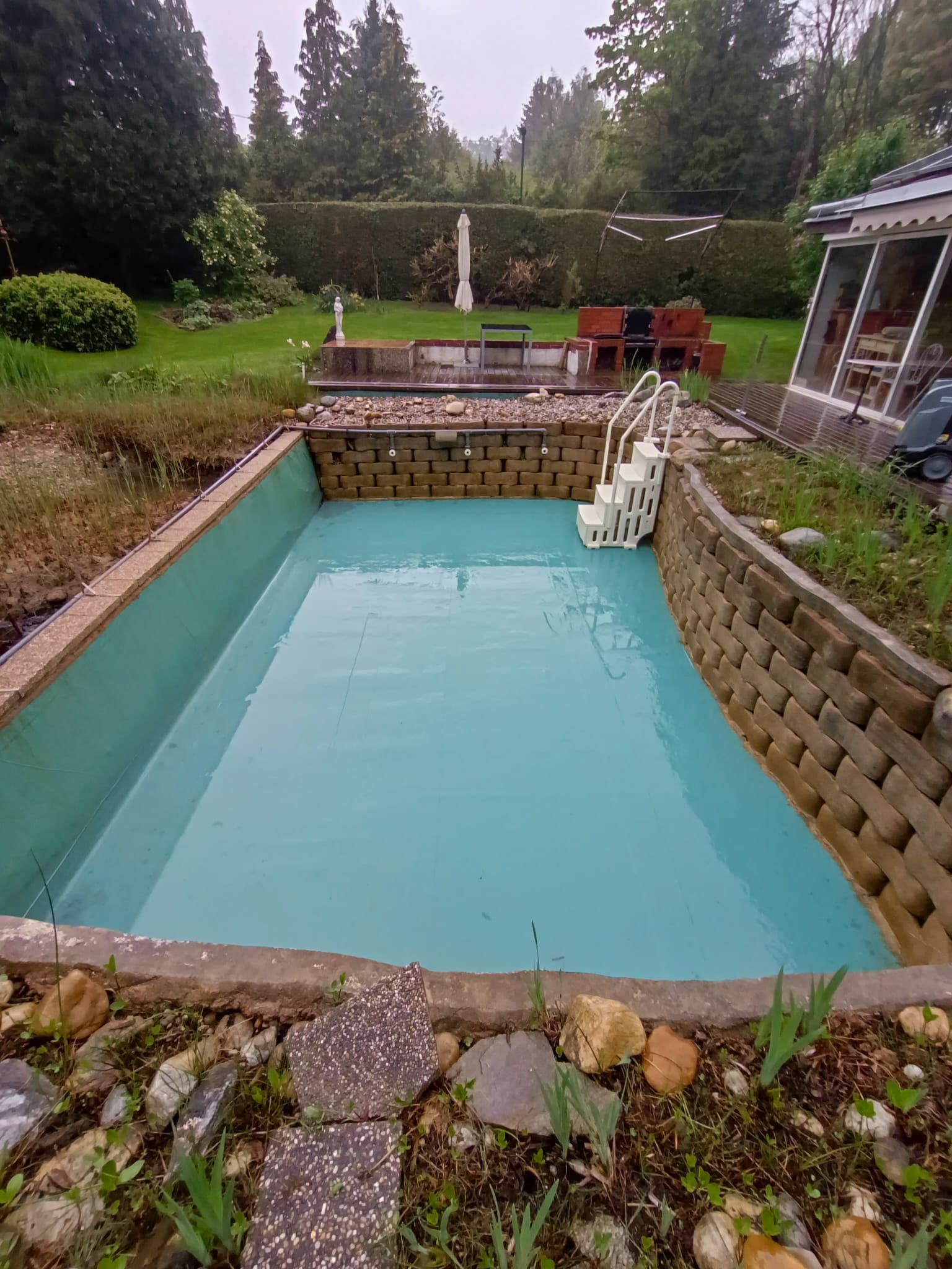 Ein leerer türkisfarbener Pool in einem Hinterhof mit Stützmauer und Treppe.