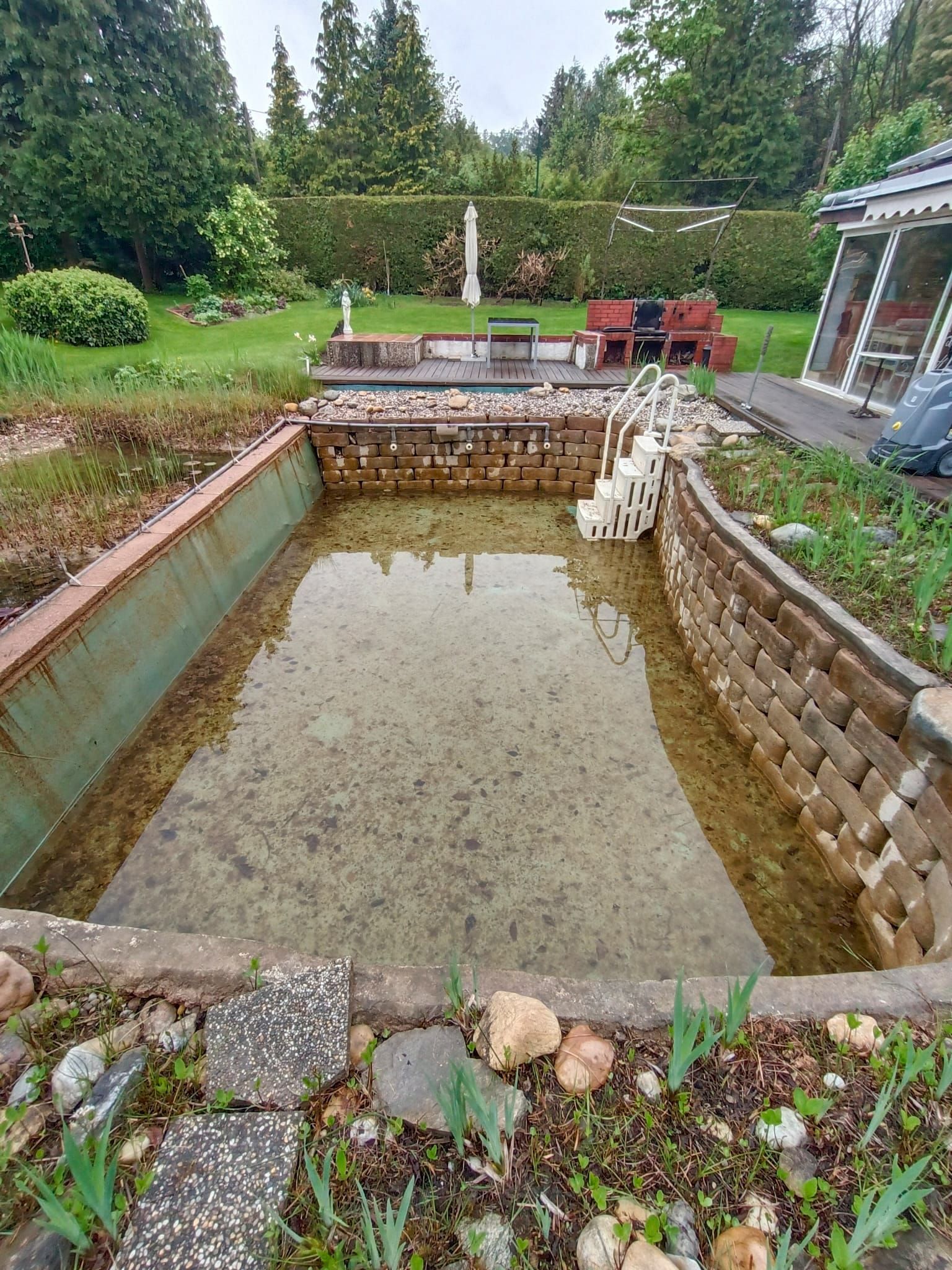 Rechteckiger, trüber Teich mit Ziegelsteineinfassung, umgeben von einem Garten und Laubwerk.