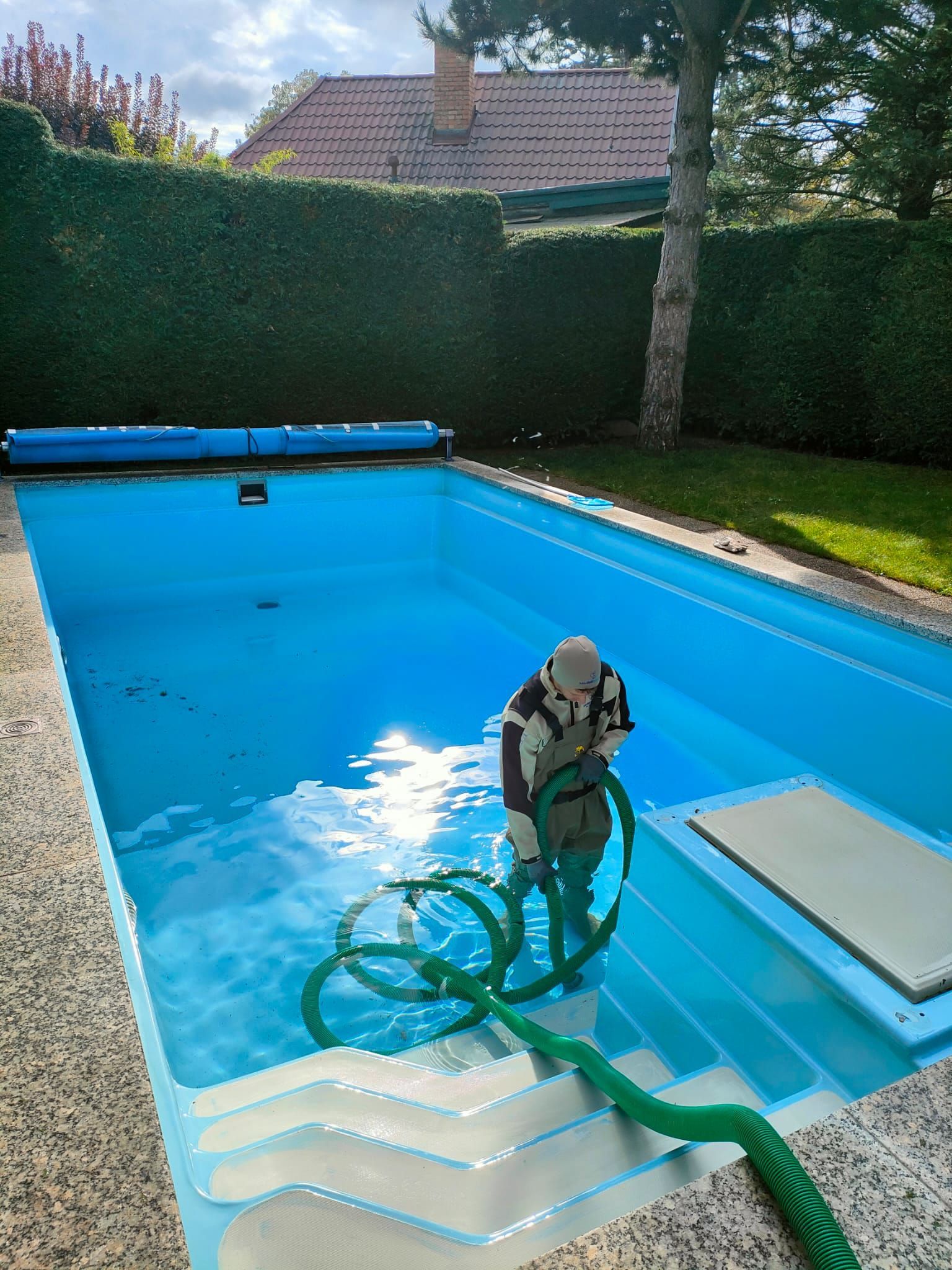 Eine Person reinigt mit einem grünen Gartenschlauch einen rechteckigen blauen Pool in einem Hinterhof.