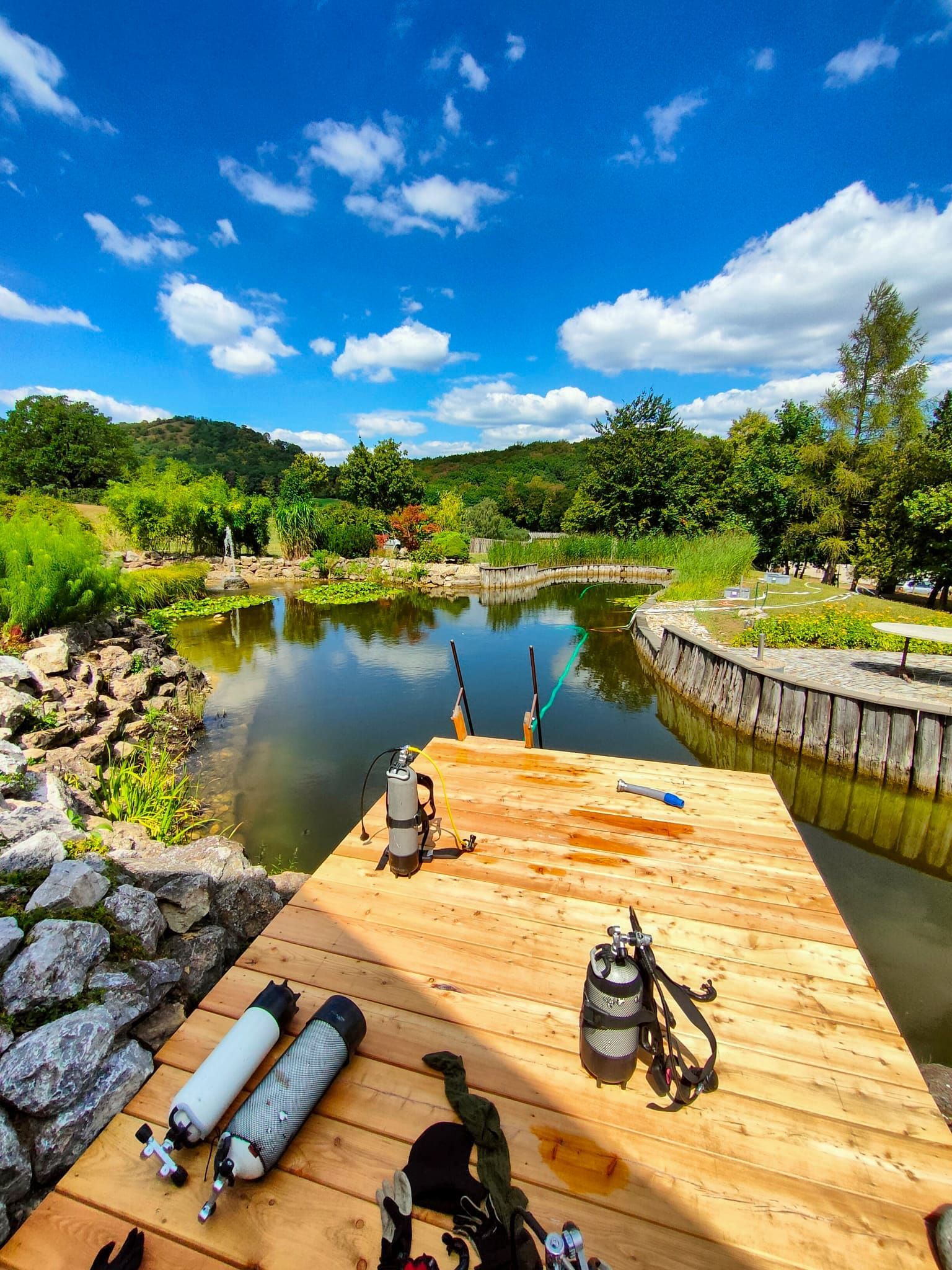 Holzsteg mit Taucherflaschen in der Nähe eines Teichs, Bäume und blauer Himmel mit Wolken.