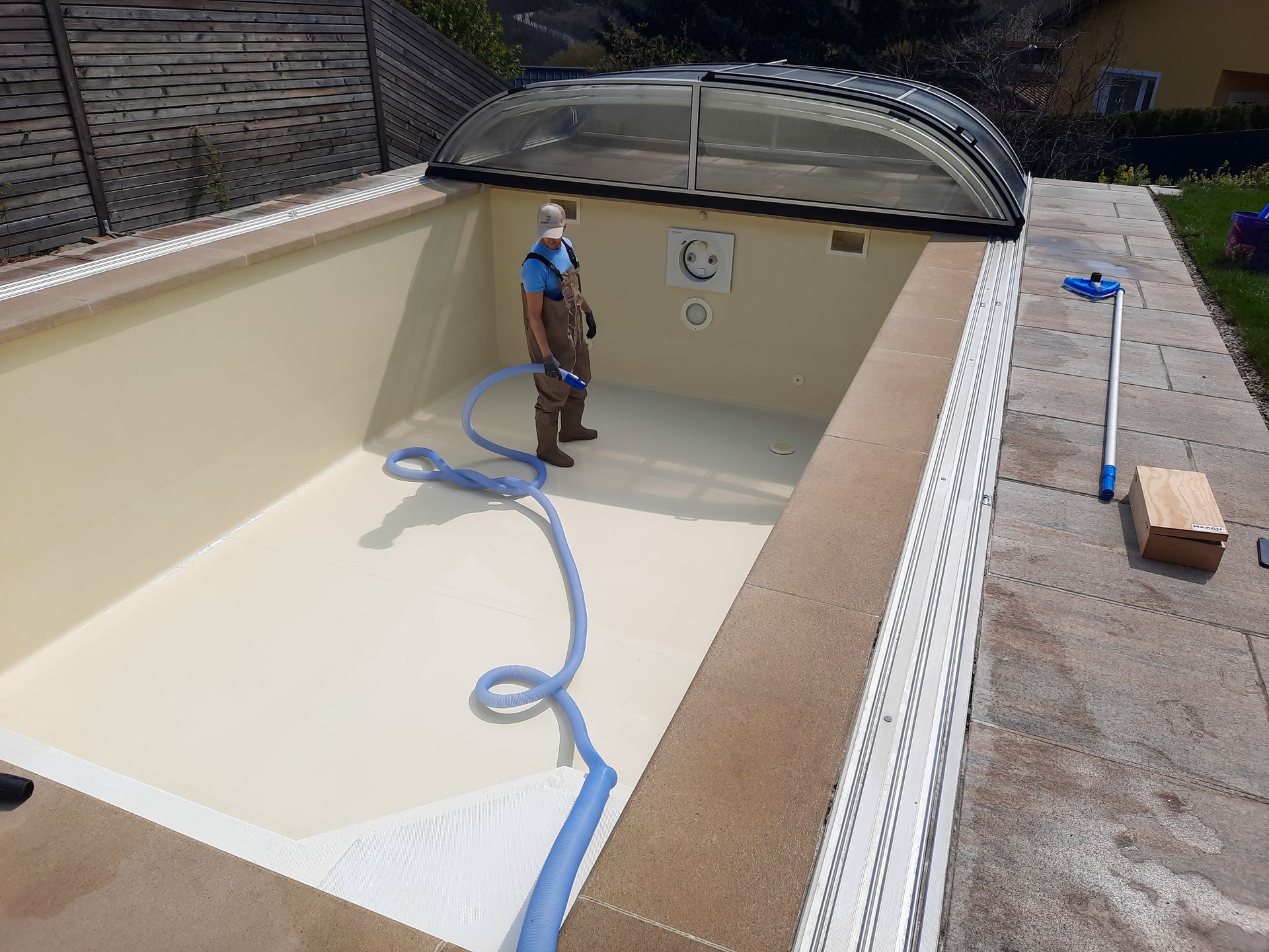 Eine Person reinigt einen leeren, beigefarbenen Pool mit einem blauen Schlauch unter einer durchsichtigen Kuppelabdeckung.