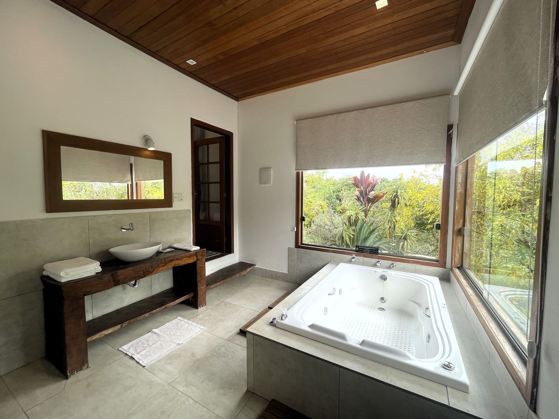 Um banheiro moderno com banheira de hidromassagem embutida ao lado de uma grande janela com vista para as árvores, e uma bancada de madeira com pia.