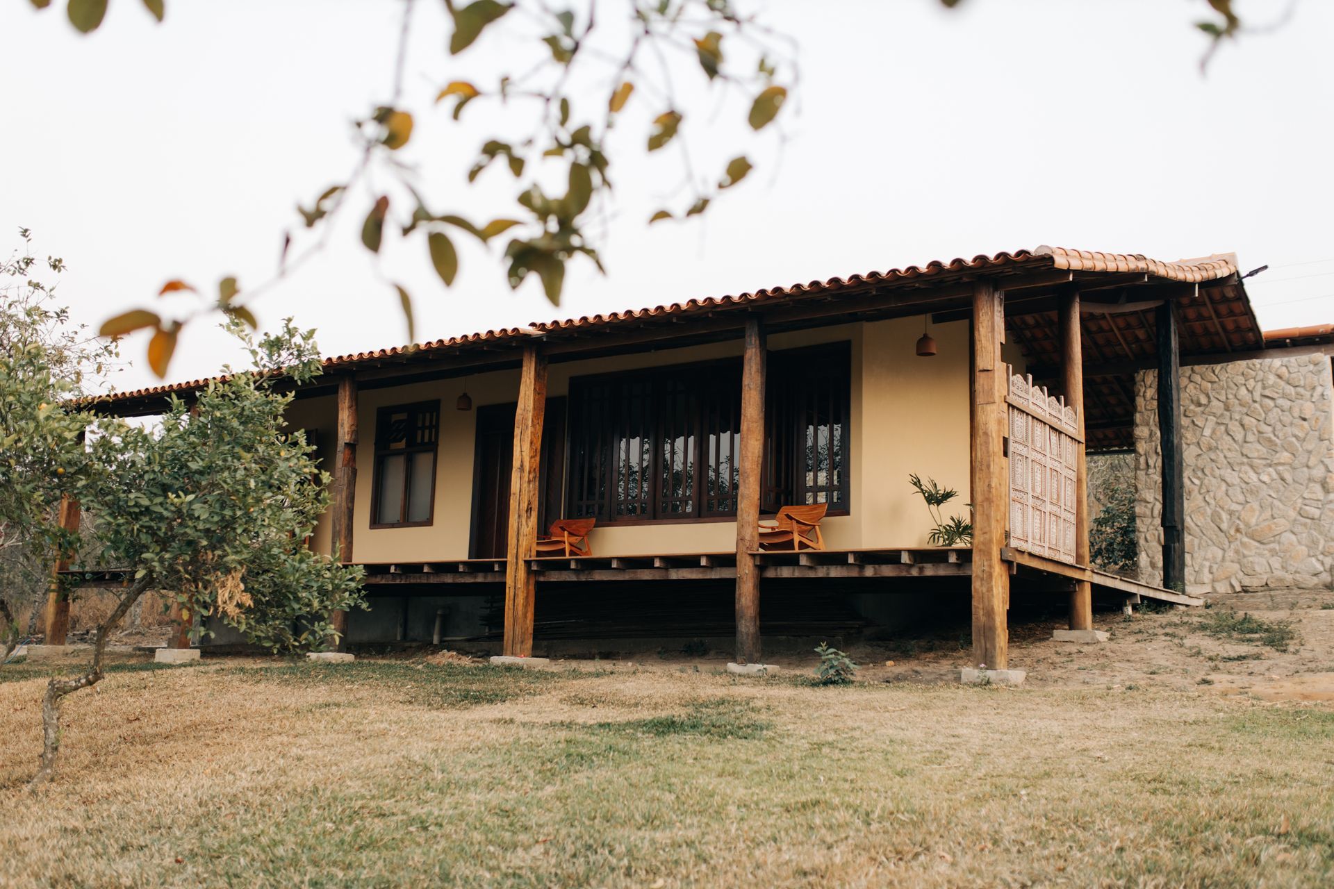 Uma cabana rústica de um só andar, com varanda de madeira e telhado de telhas, situada em uma colina gramada sob um céu nublado.