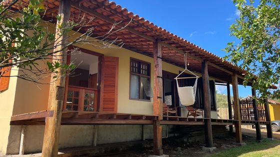 Uma casa bege com um amplo deck de madeira e cobertura na varanda, com uma cadeira suspensa sob um céu azul brilhante e ensolarado.