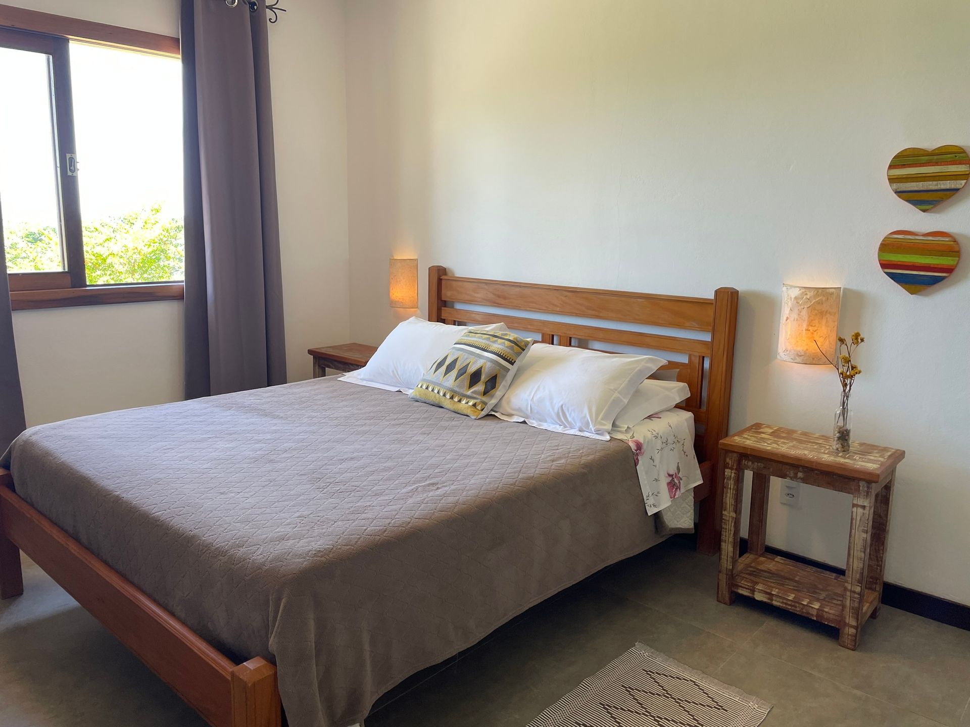 Um quarto simples com uma cama de madeira, colcha marrom, dois travesseiros brancos, mesas de cabeceira com abajures e decoração de coração na parede.