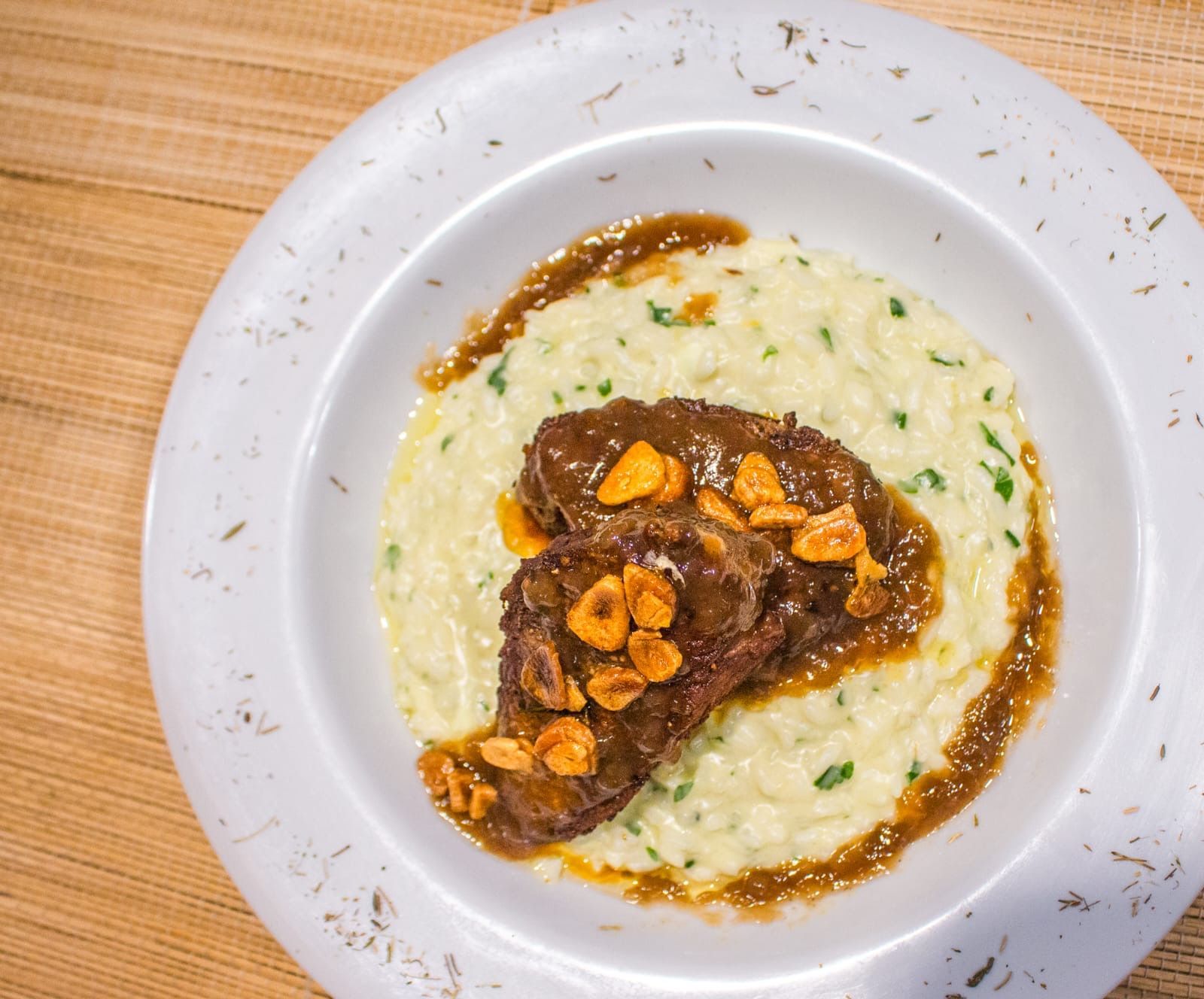 Um prato de carne assada lentamente, coberta com lascas de alho crocantes, servida sobre uma cama de risoto cremoso com ervas, em uma tigela branca.