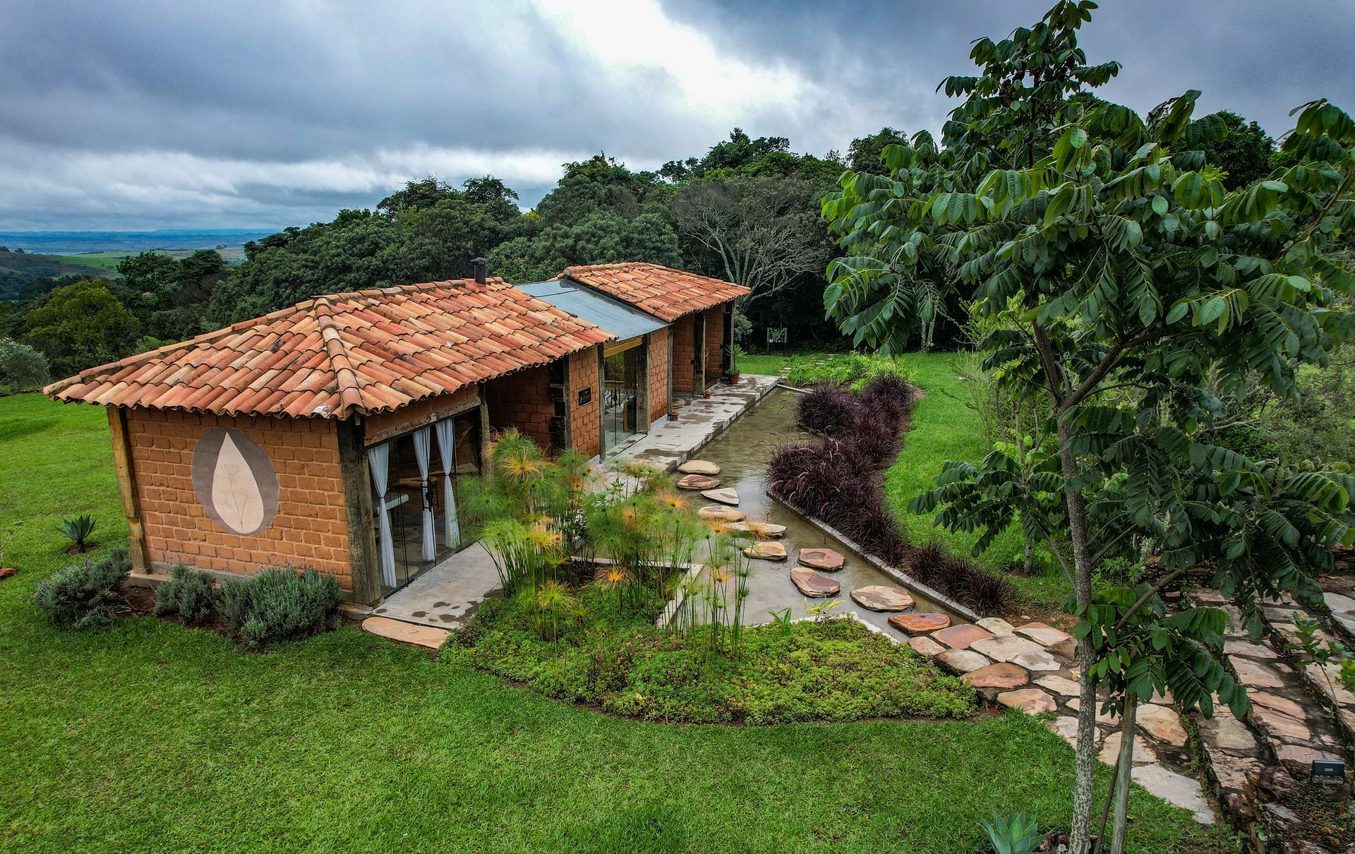 Uma construção rústica de tijolos com telhado de telhas fica em uma colina gramada, com um caminho de pedra que passa por ela, atravessando um jardim.