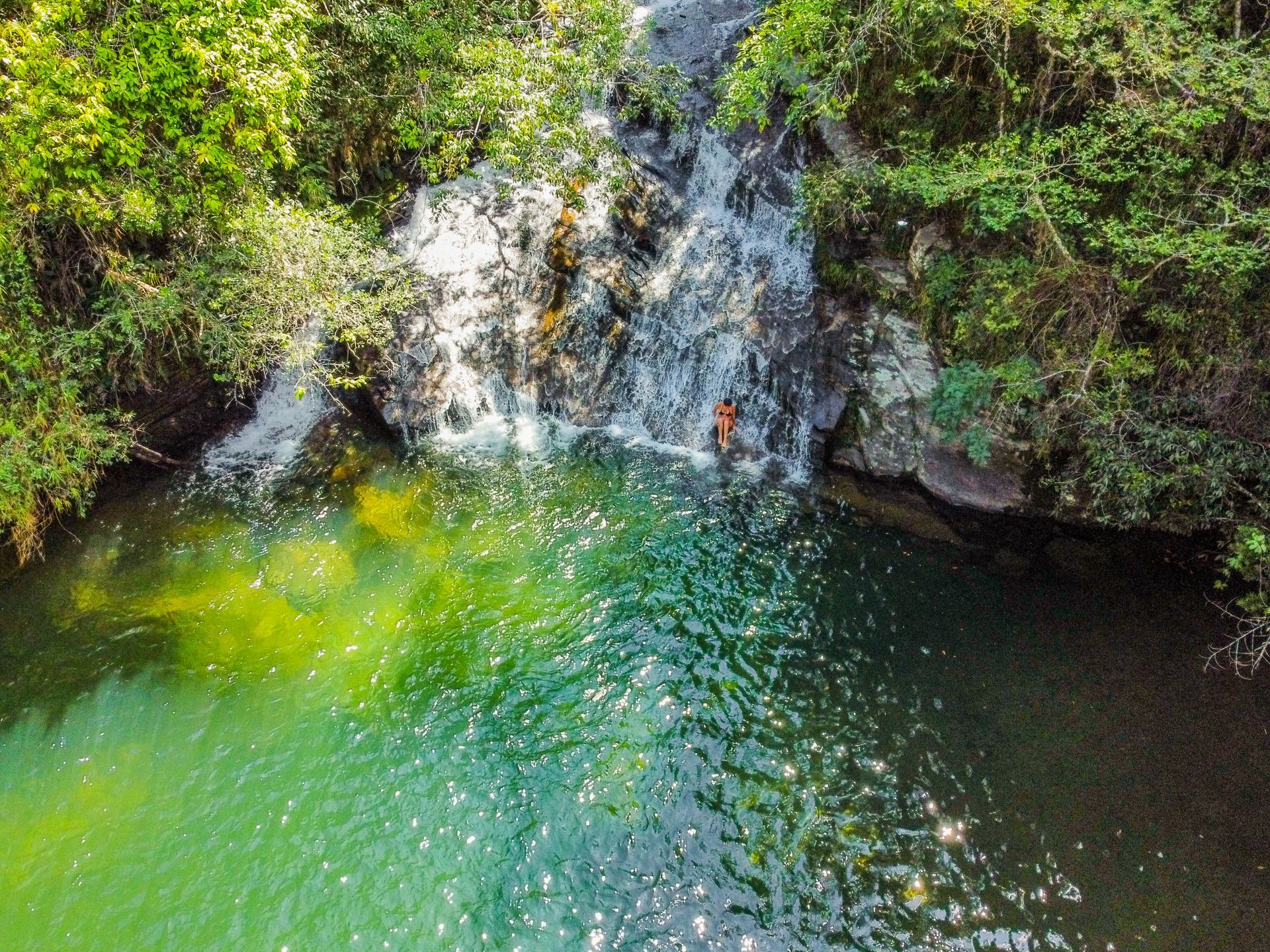 Uma pessoa está em pé em uma cachoeira rochosa que deságua em uma piscina natural de água verde cristalina, cercada por uma floresta exuberante.