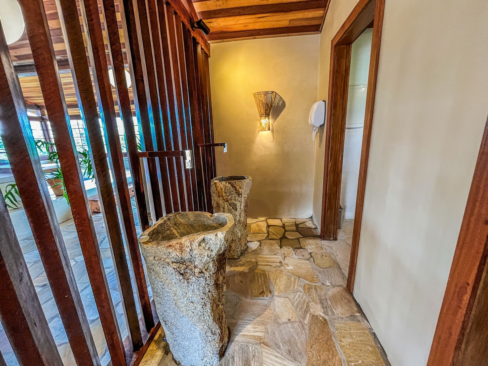 Duas pias rústicas de pedra com pedestal em um cômodo revestido com azulejos, paredes com ripas de madeira e iluminação aconchegante.