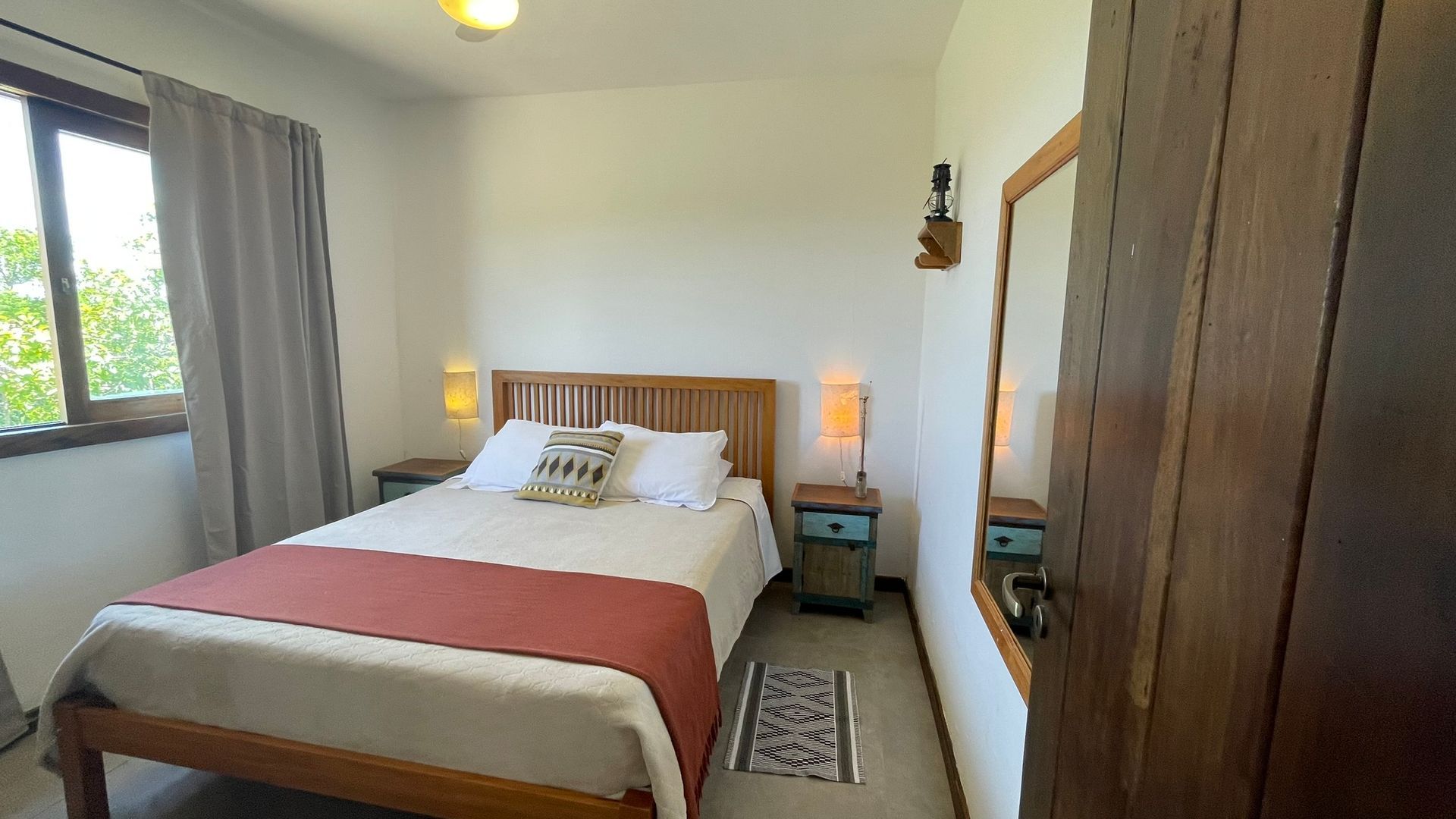 Um quarto com uma cama de casal queen-size, com uma peseira cor de ferrugem, cabeceira de madeira, mesas de cabeceira, luminárias e um espelho de parede.