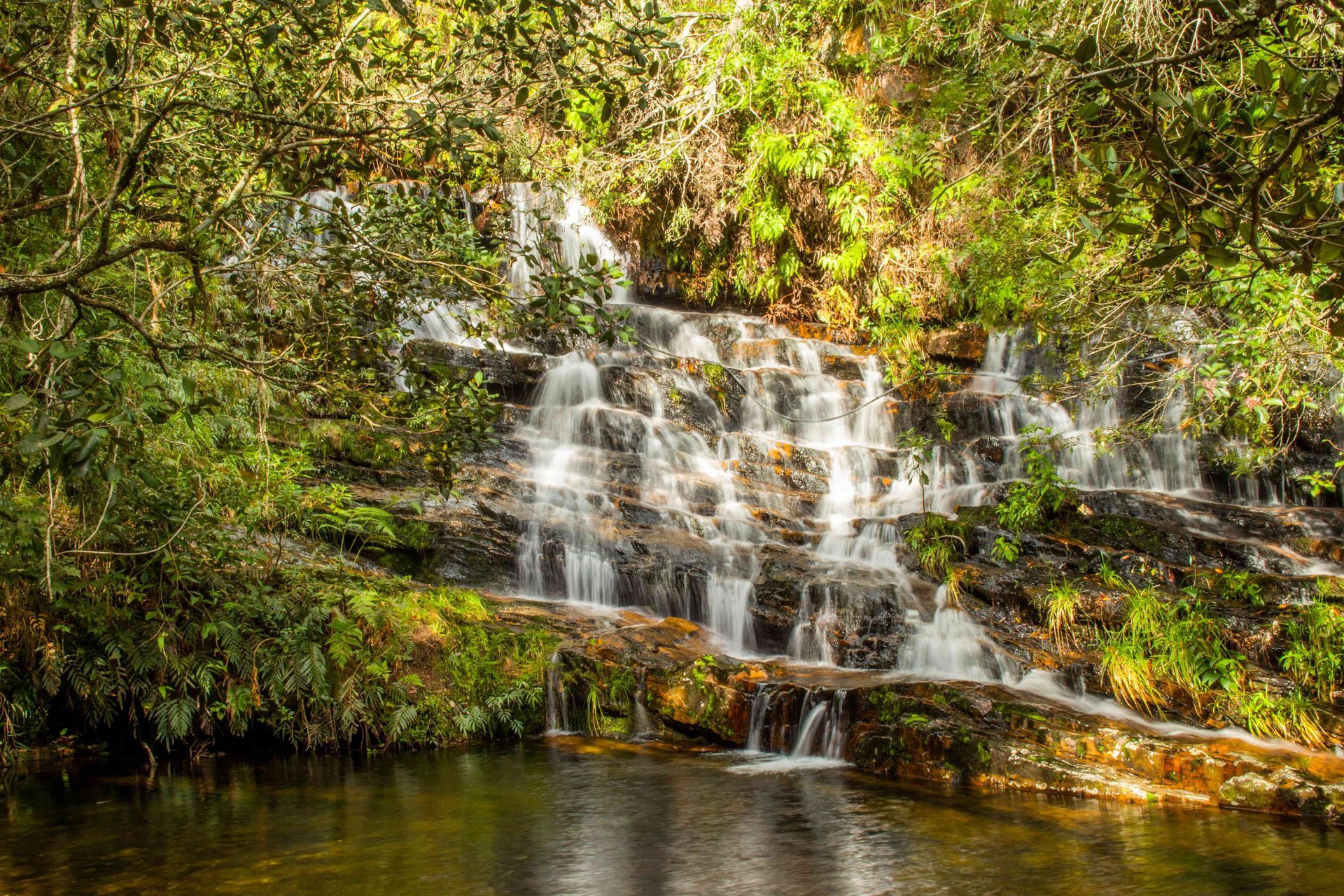 Uma cascata de vários níveis despenca sobre rochas escuras em uma piscina, cercada por folhagem verdejante em uma floresta ensolarada.
