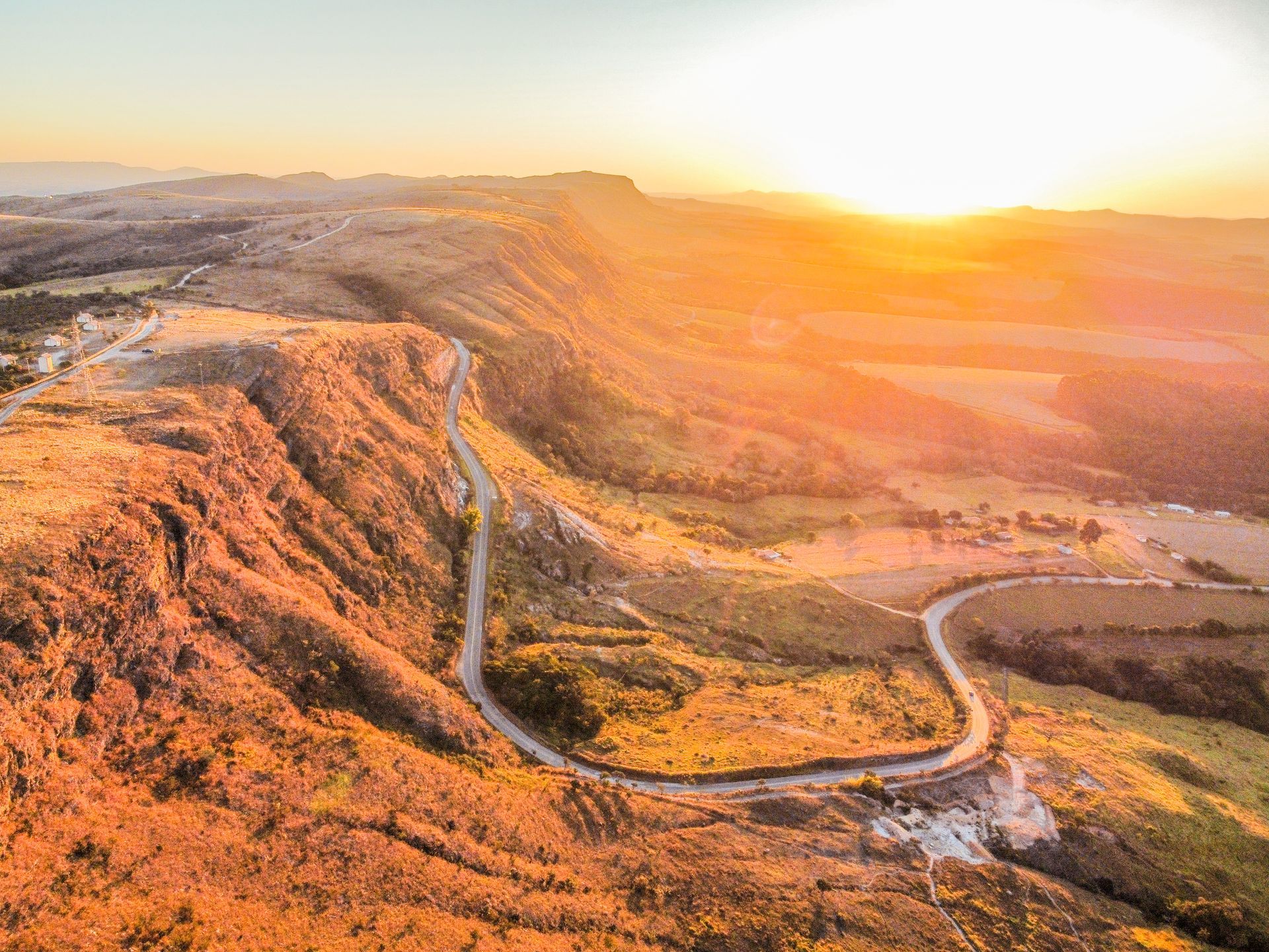 Ao pôr do sol, uma estrada sinuosa serpenteia pela beira de um penhasco acidentado, lançando um brilho dourado e quente sobre a paisagem montanhosa.