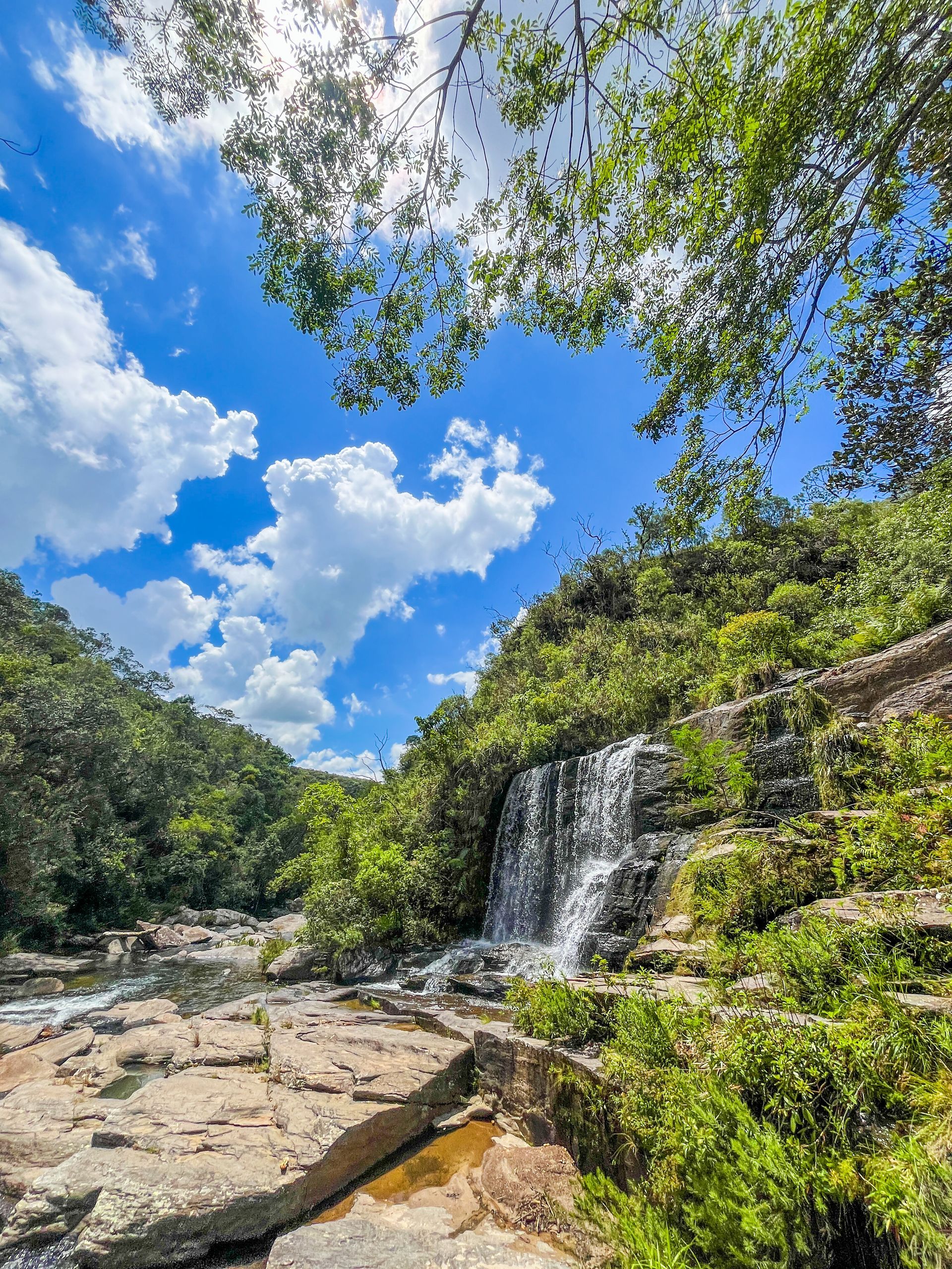 Uma pequena cachoeira que deságua em um riacho, sobre rochas em camadas, cercada por árvores verdejantes sob um céu azul brilhante.