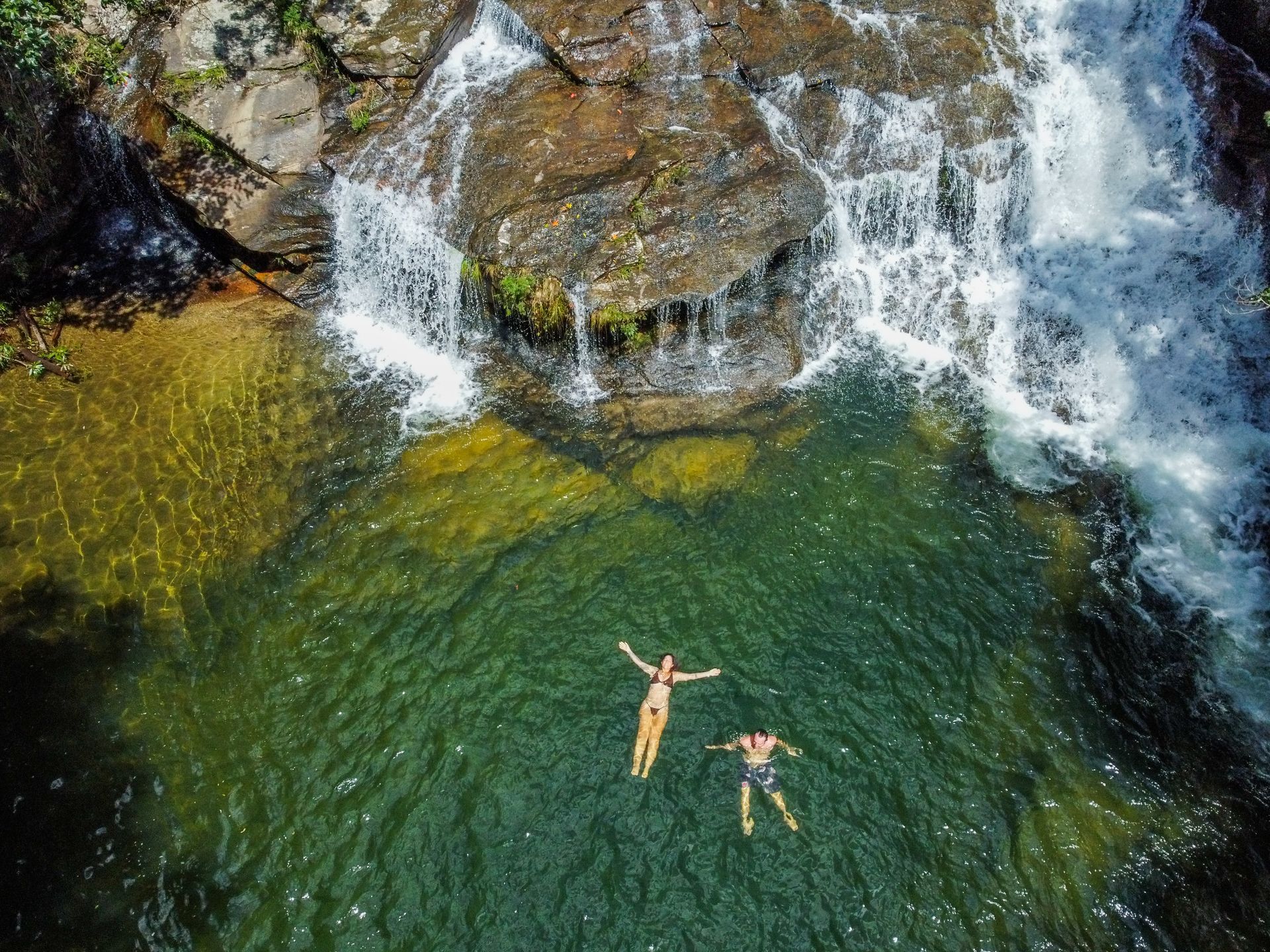 Duas pessoas flutuam em uma piscina verde e cristalina na base de uma pequena cachoeira, cercada por terreno rochoso.