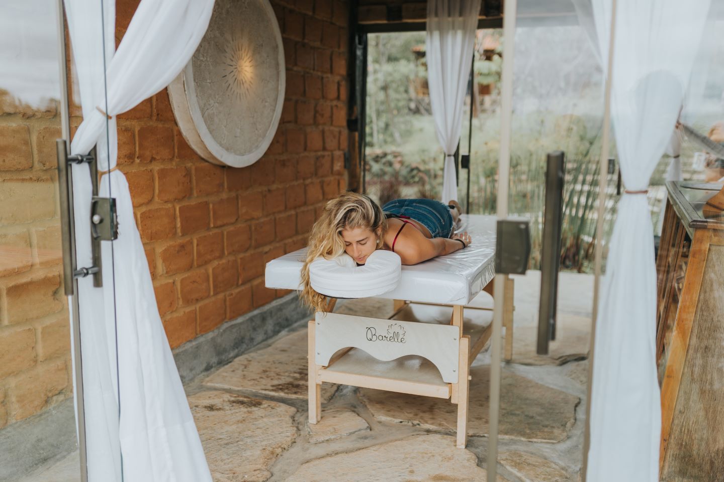 Uma pessoa está deitada de bruços em uma mesa de massagem em um pavilhão ao ar livre com paredes de pedra, cortinas brancas e luz natural.