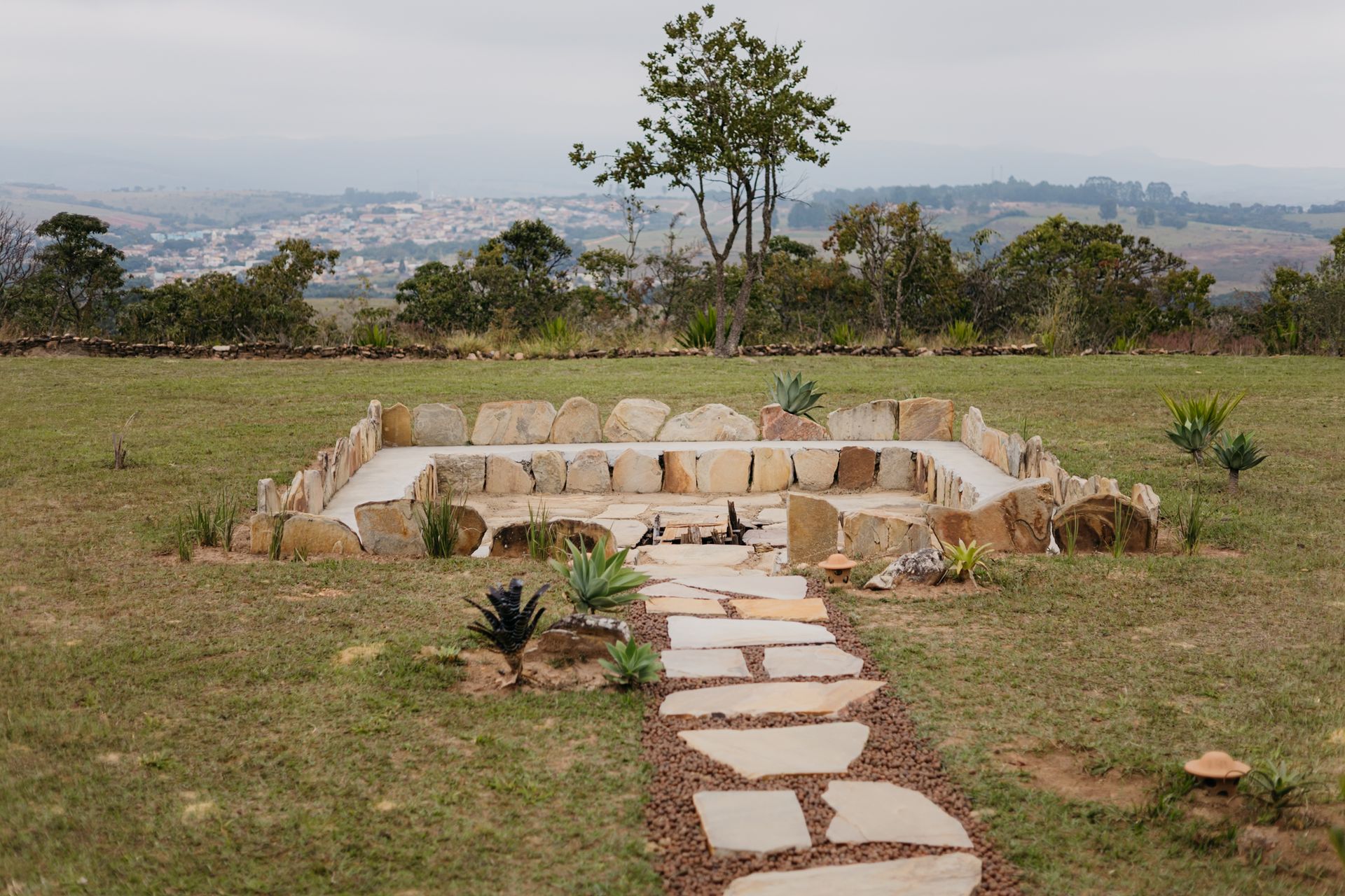 Um caminho pavimentado com pedras leva a uma área retangular com uma fogueira de pedra em um campo gramado, com uma cidade ao fundo.