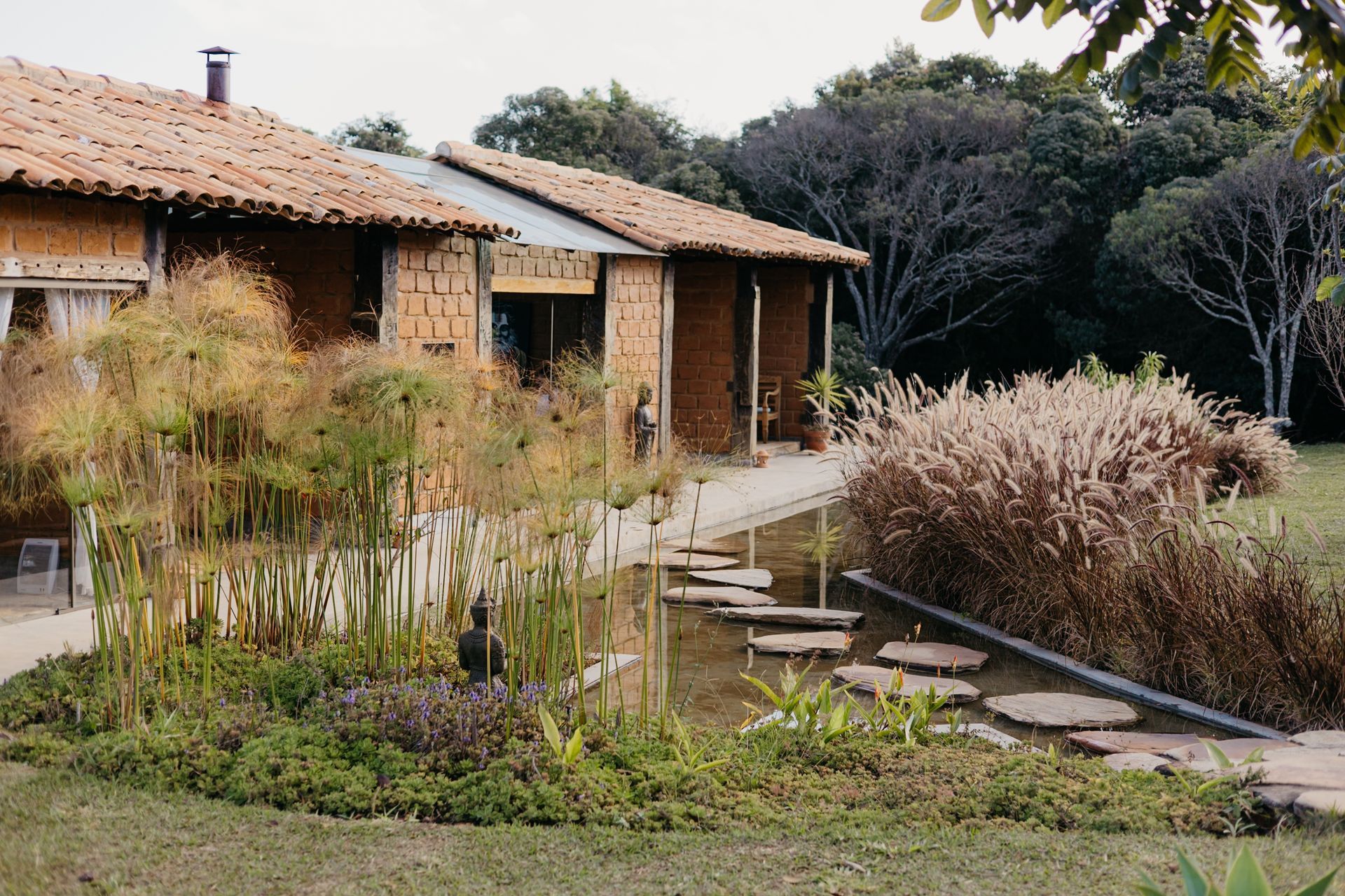 Uma construção rústica, com telhas de terracota e paredes de tijolos de barro, tem vista para um lago com pedras para atravessar e grama alta.