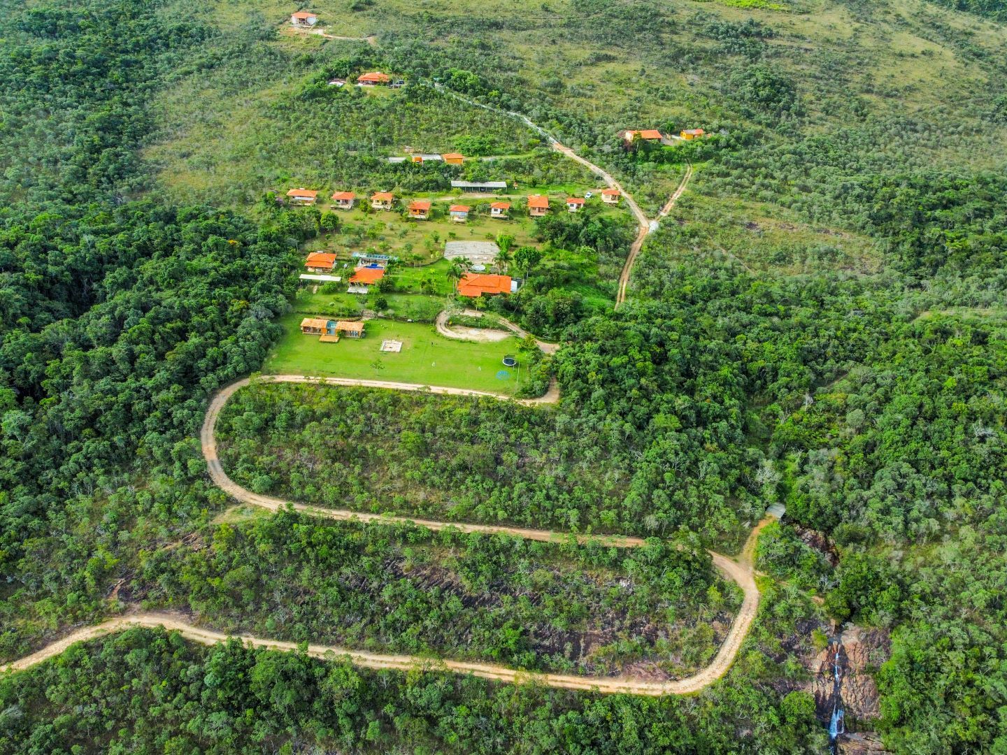 Vista aérea de um pequeno conjunto de edifícios com telhados alaranjados, cercado por floresta, acessível por uma estrada de terra sinuosa.