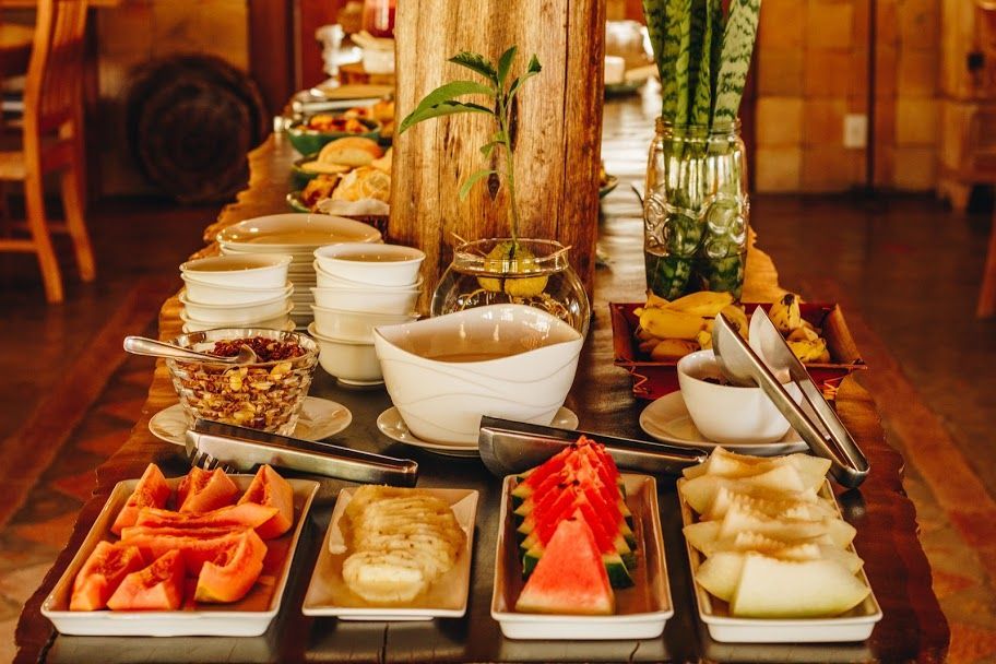 Uma mesa de buffet de café da manhã com pratos de frutas fatiadas, tigelas pequenas, pilhas de louça e um vaso de folhas verdes.