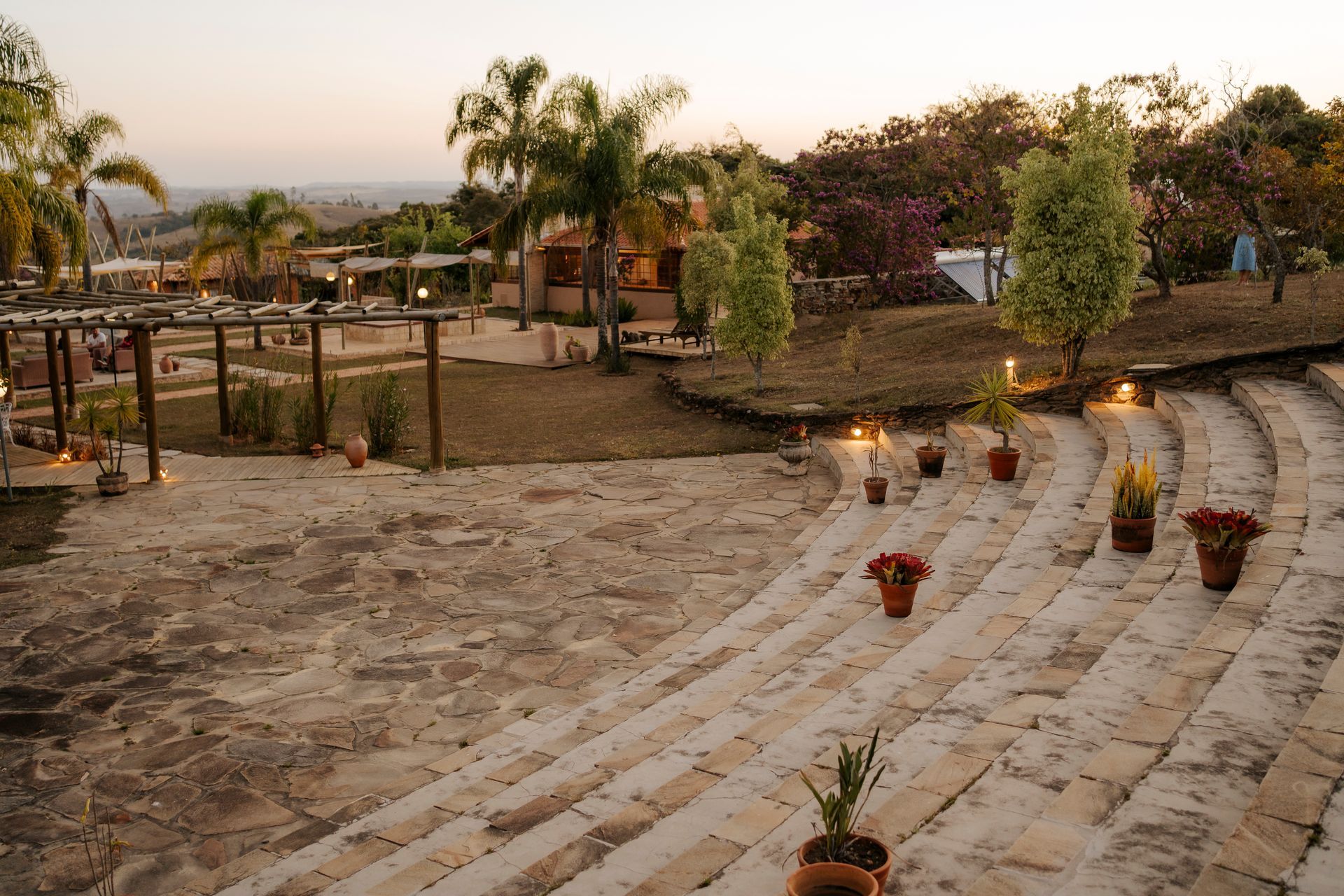 Degraus de pedra conduzem a uma área externa com palmeiras e um edifício ao entardecer, realçado por vasos de plantas e iluminação suave.