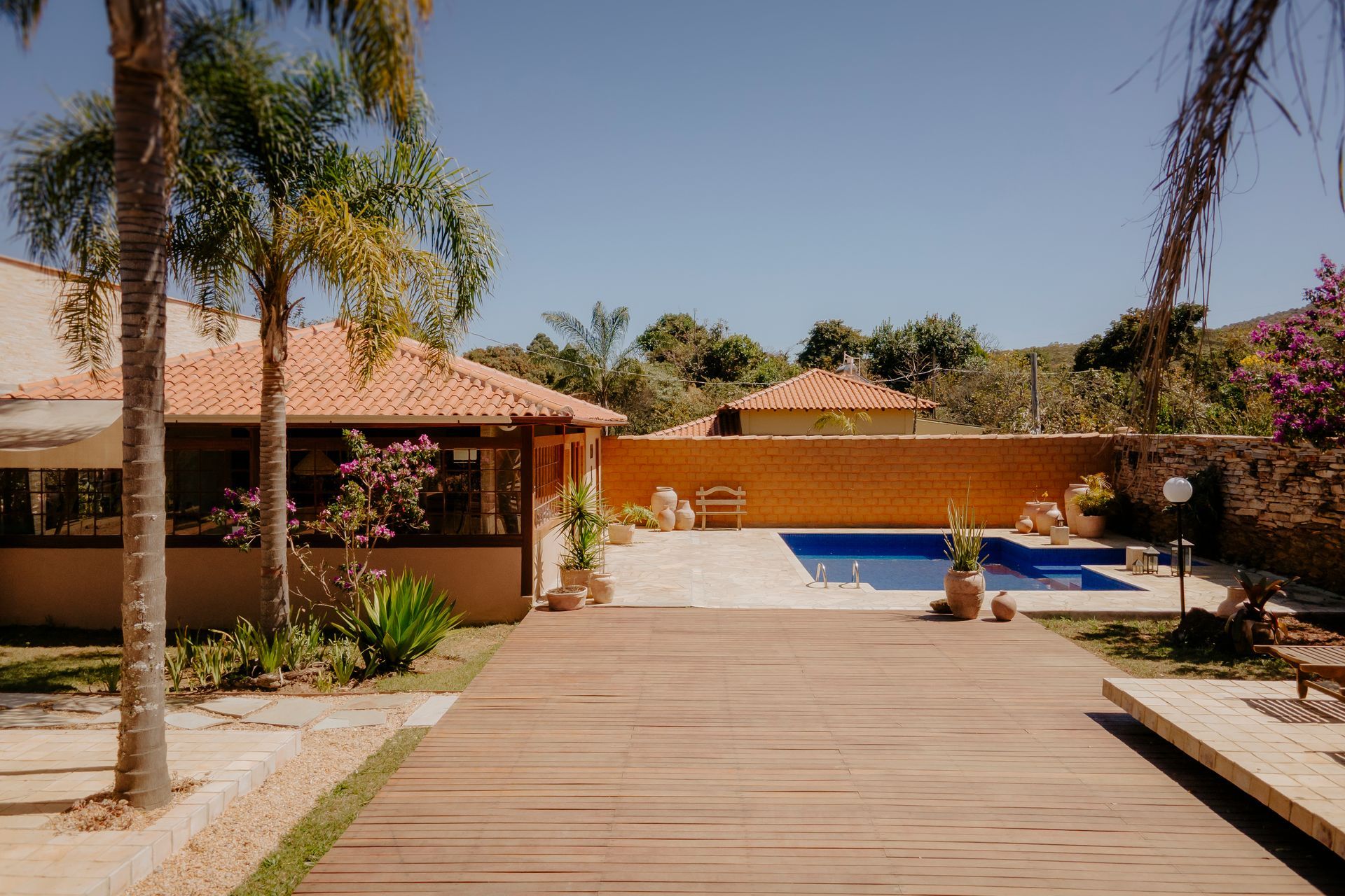 Um deck de madeira conduz a uma piscina ao lado de uma casa com telhado de terracota, palmeiras e uma parede de tijolos amarelos.