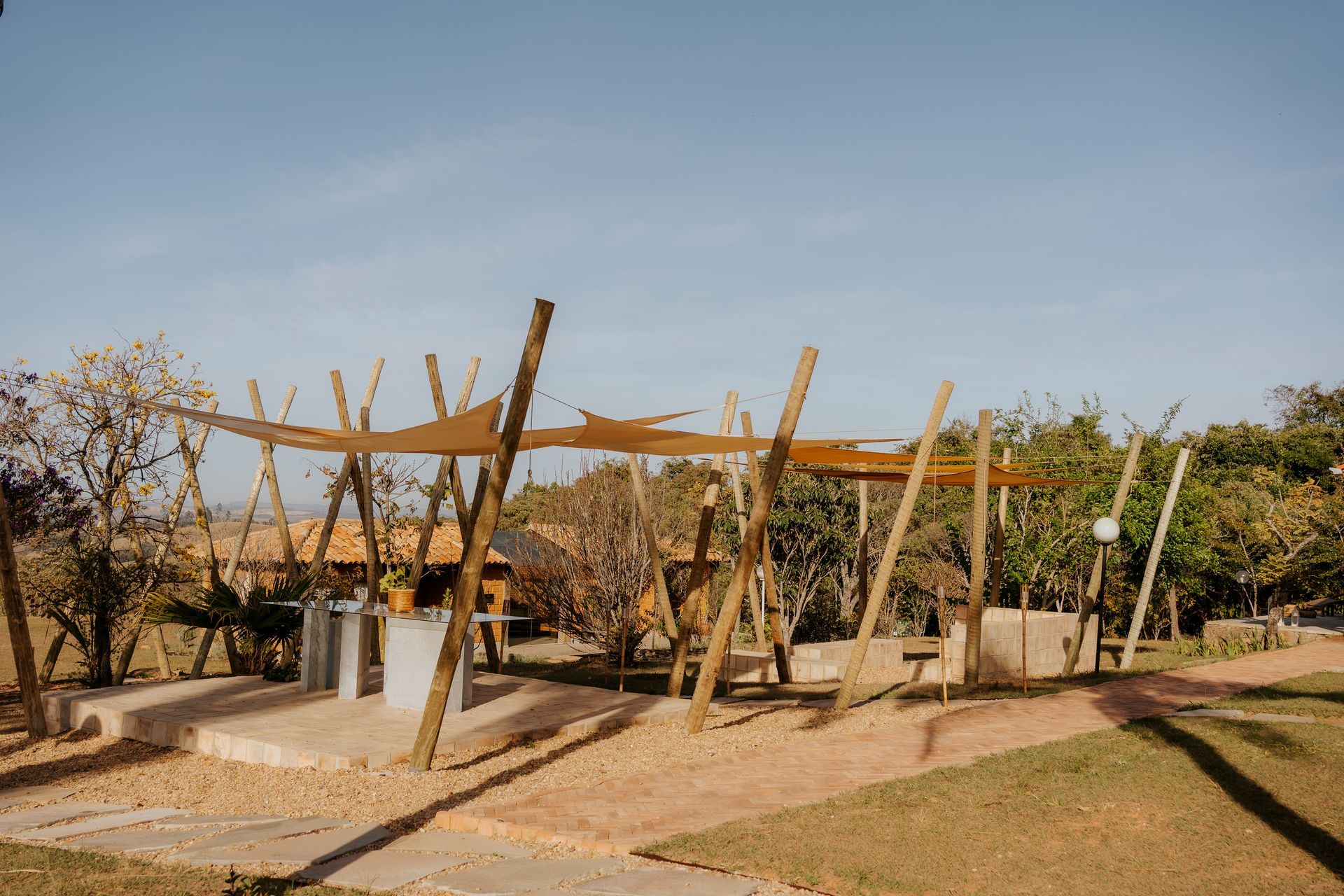 Uma área de pátio externa com uma cobertura de tecido bege sustentada por postes de madeira, situada em um terraço de cascalho próximo a árvores.