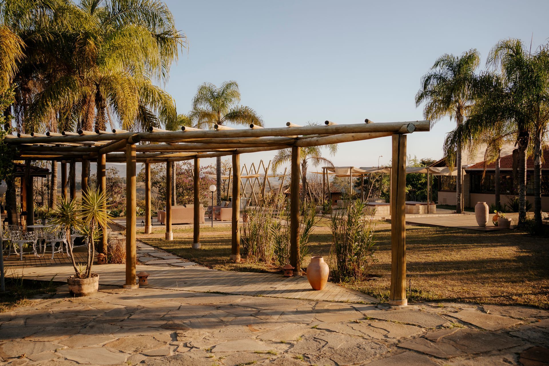 Uma pérgola de madeira sombreia um pátio de pedra rodeado por palmeiras e vegetação típica do deserto, sob um céu claro e ensolarado.