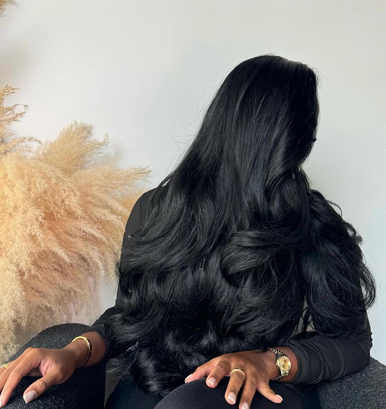 Mujer con cabello largo y ondulado de color negro, vestida con una camisa negra de manga larga y joyas de oro, sentada.