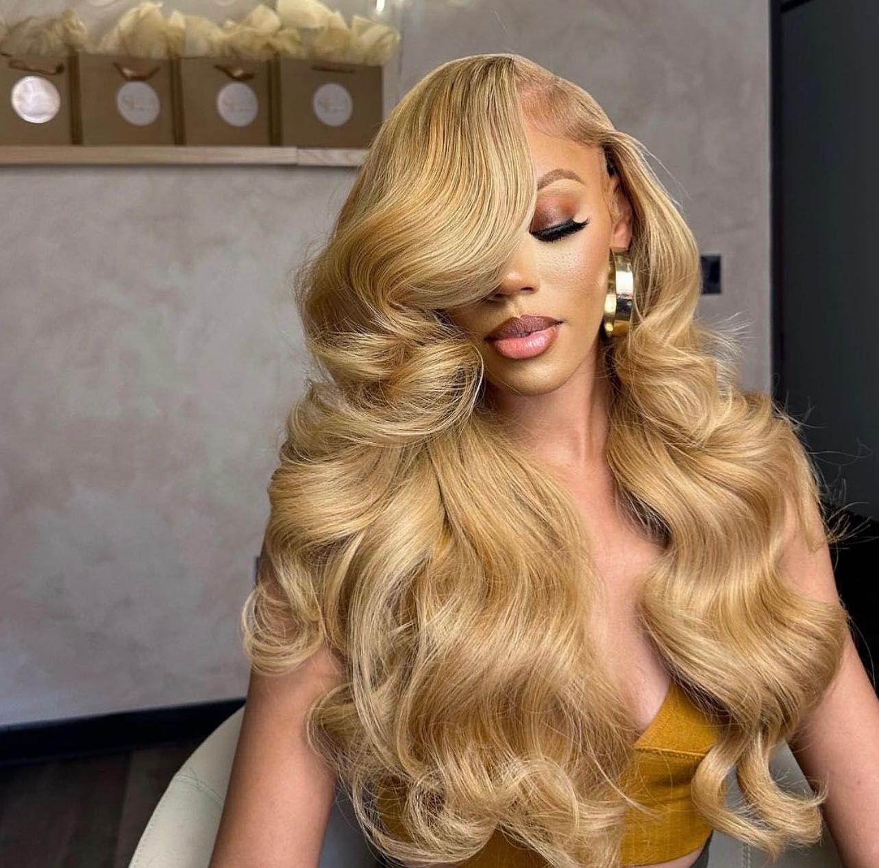 Mujer con cabello rubio largo y ondulado peinado hacia un lado, con grandes aros dorados y un top amarillo.