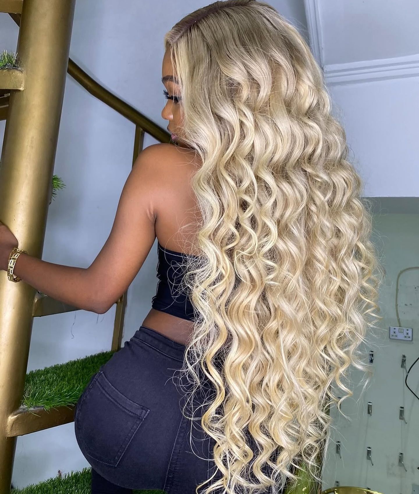 Mujer con cabello rubio largo y ondulado, vestida con un top negro y jeans, posó cerca de una escalera de caracol dorada.