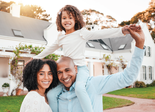 Family of three smiling in front of a house. Child on father's shoulders. Sunny day.