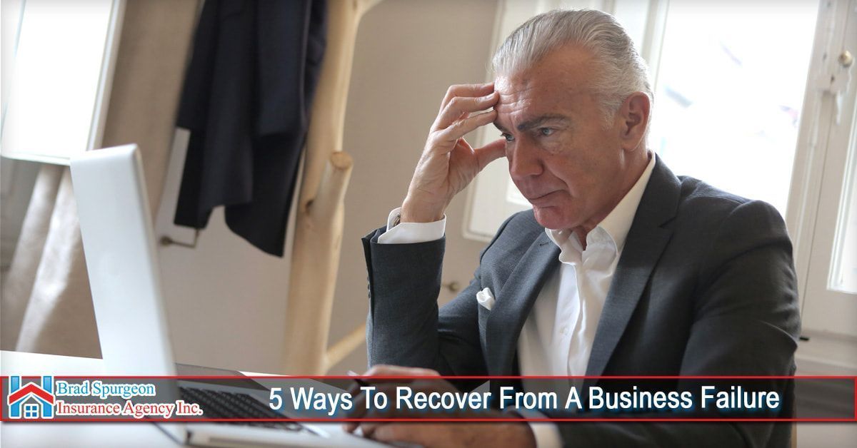 A person in a suit sits at a laptop looking stressed with their hand to their forehead.
