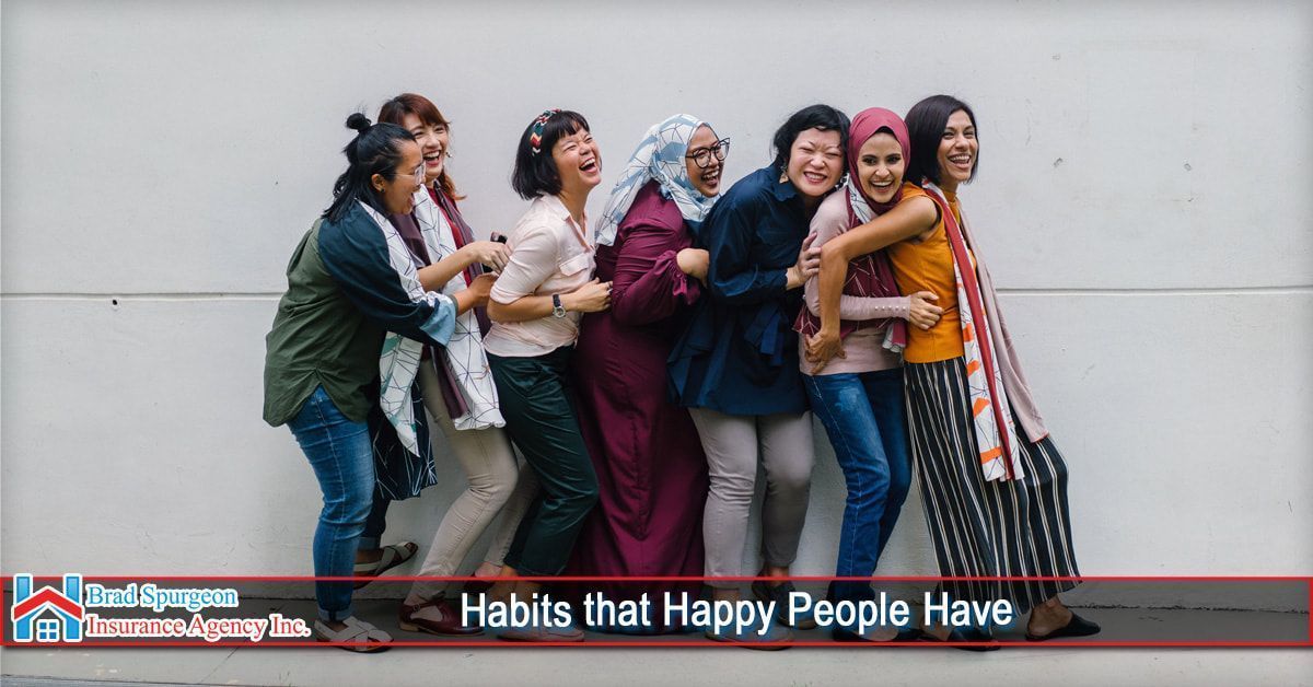 A group of smiling people stand in a line, hugging each other against a plain wall. 