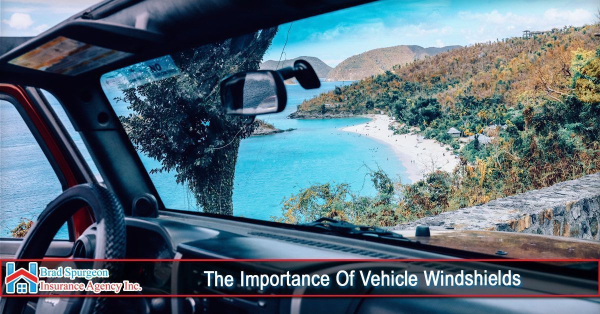 View from inside a vehicle through a windshield overlooking a scenic tropical beach with blue water and hills.
