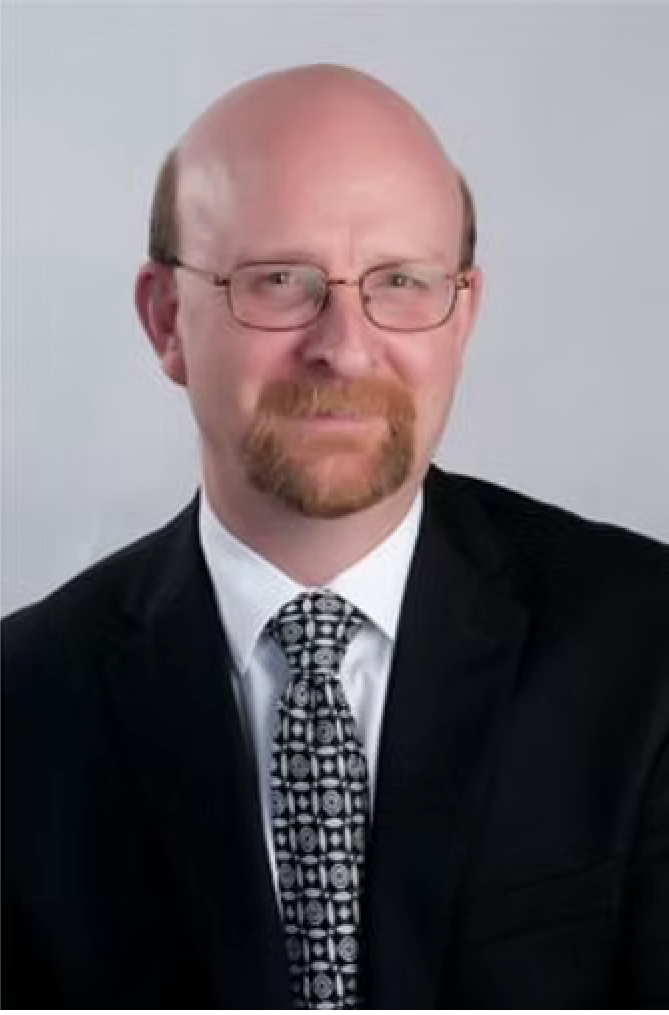 A portrait of a smiling person with dark, wavy hair, wearing a gray shirt and dark jacket against a textured wood backdrop.
