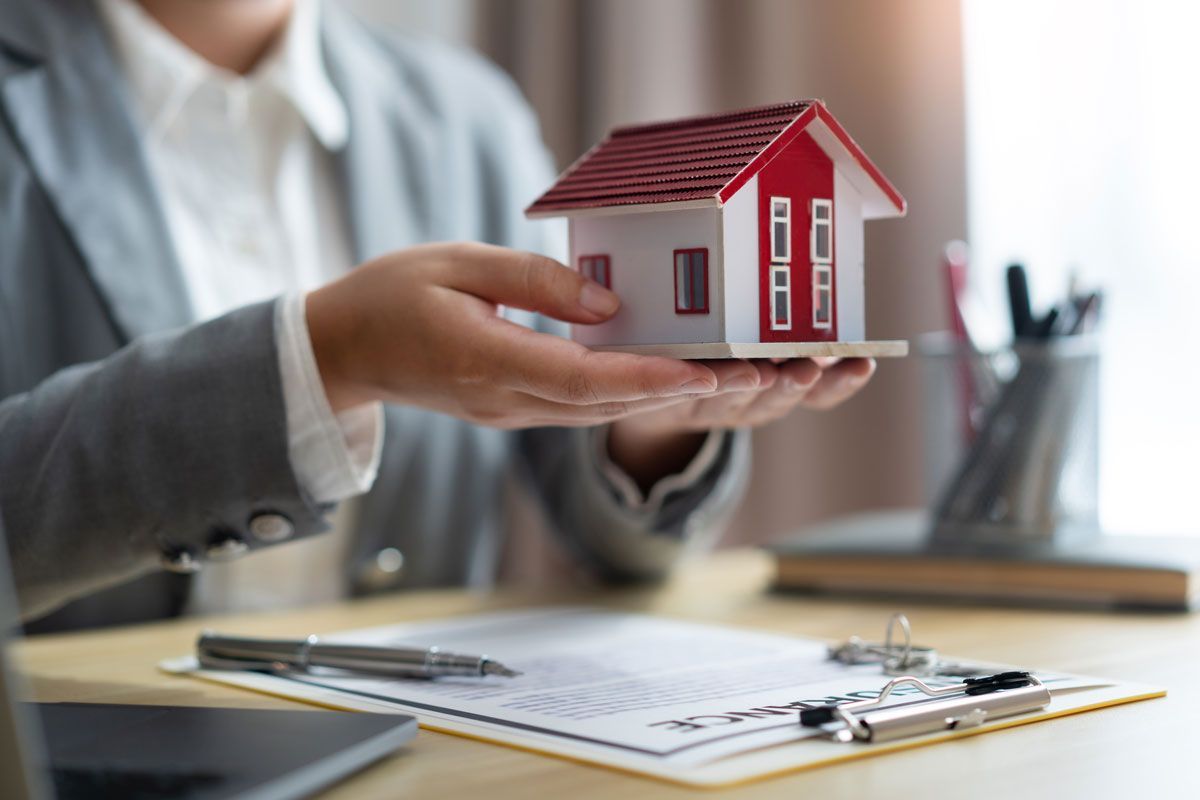 A person in professional attire holds a small model house above a desk with documents and pens.