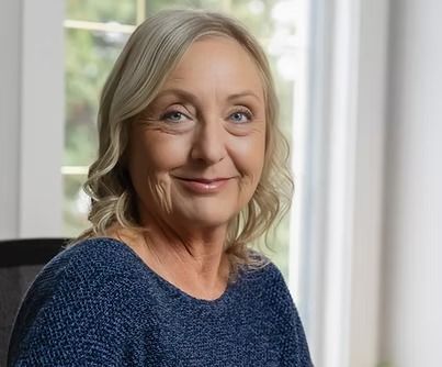 A smiling person with blonde hair wearing a blue textured top, positioned indoors near a window.
