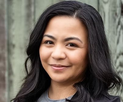 A person with shoulder-length dark hair smiling gently, captured in a headshot against a textured, light-colored backdrop.