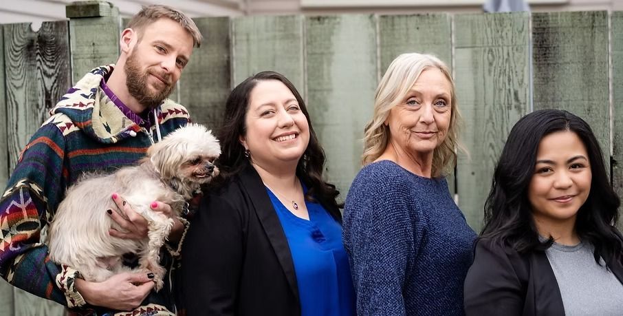 Four people stand together in front of a wood-paneled fence; one person holds a small dog in their arms.