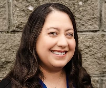 A person with dark, wavy hair smiles warmly in front of a textured gray stone wall.