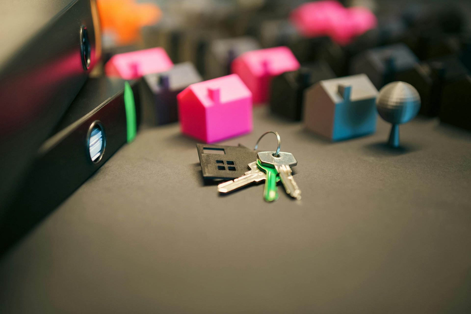 A set of metal keys rests on a gray surface surrounded by small, colorful 3D-printed model houses and black binders.