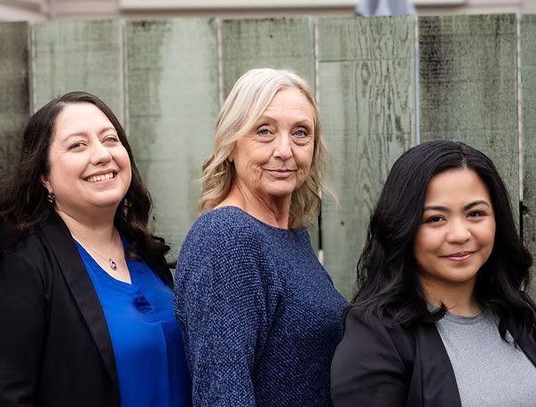Three women in front of a wooden fence; smiling. One in a blue shirt, the others in dark jackets.
