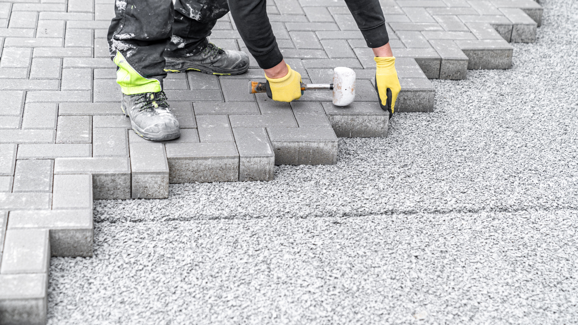 Person laying gray paving stones with a mallet on gravel, outdoors.