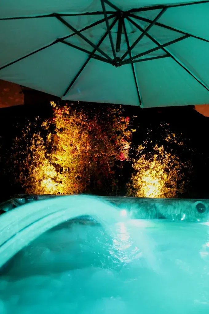 Hot tub at night with teal water, under an umbrella, illuminated by golden lights in background.