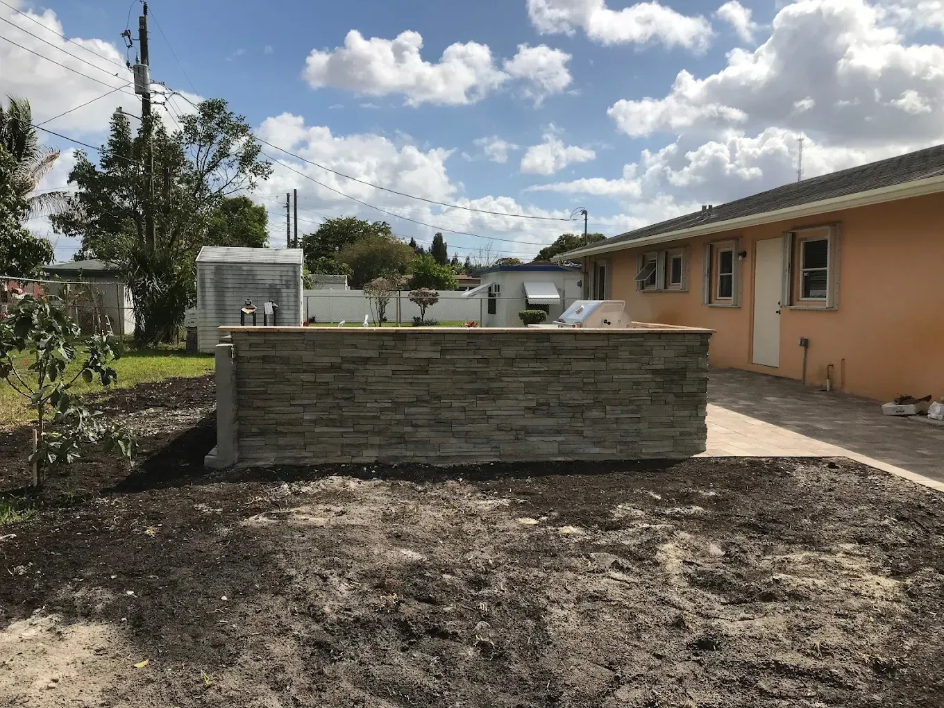 Stone wall in front of a house, set in a yard with recently tilled soil, under a cloudy sky.