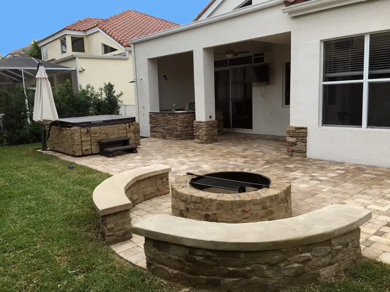 Backyard patio with fire pit, stone seating, hot tub, and covered area; beige brick and greenery.