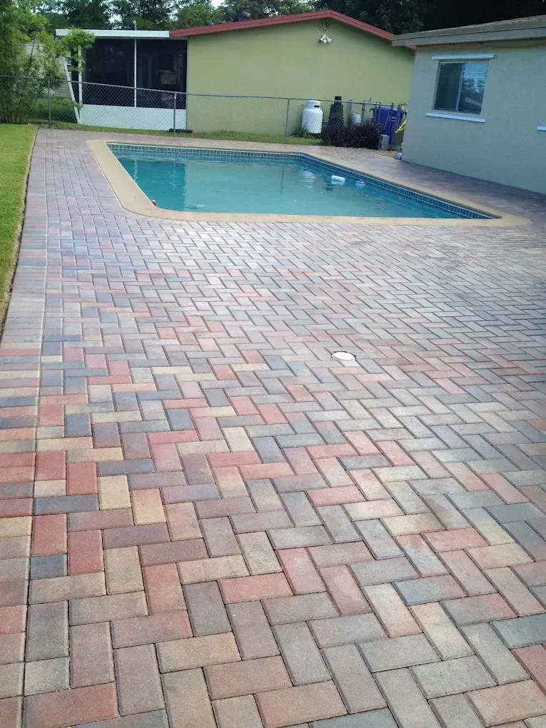Brick patio surrounds a blue pool in a backyard. A green building is visible behind the pool.