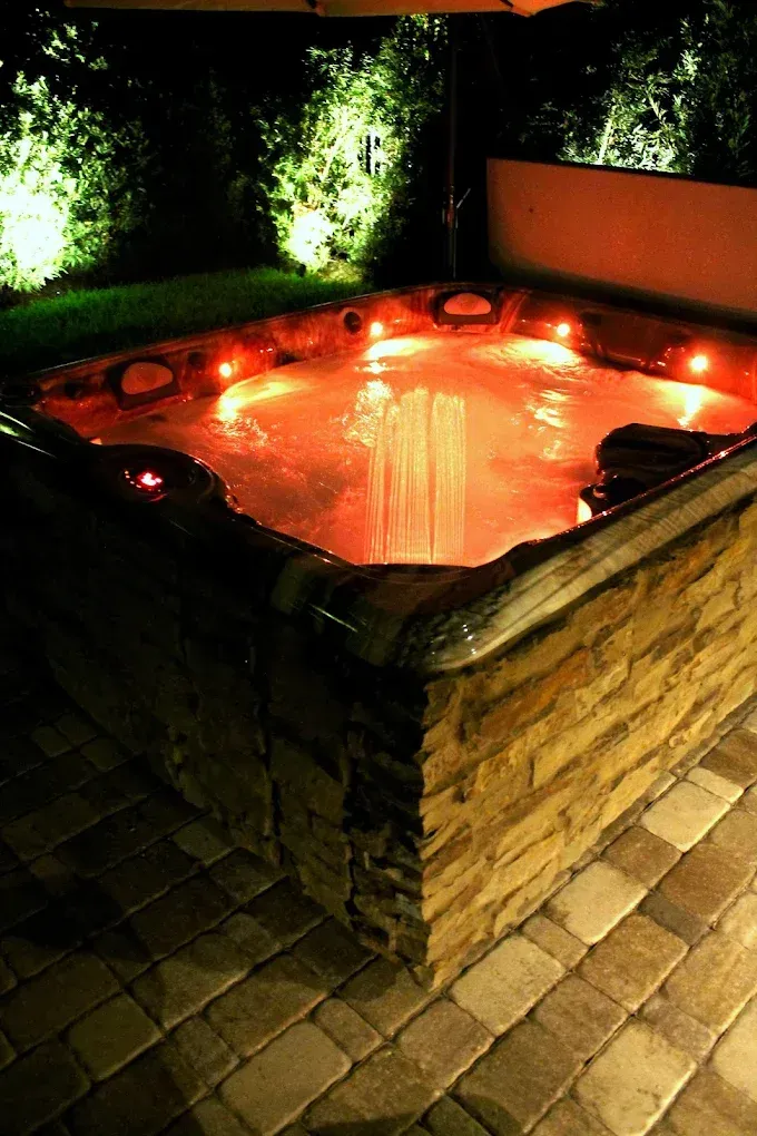 Lit hot tub at night with brick surround and illuminated jets, set in a patio area.