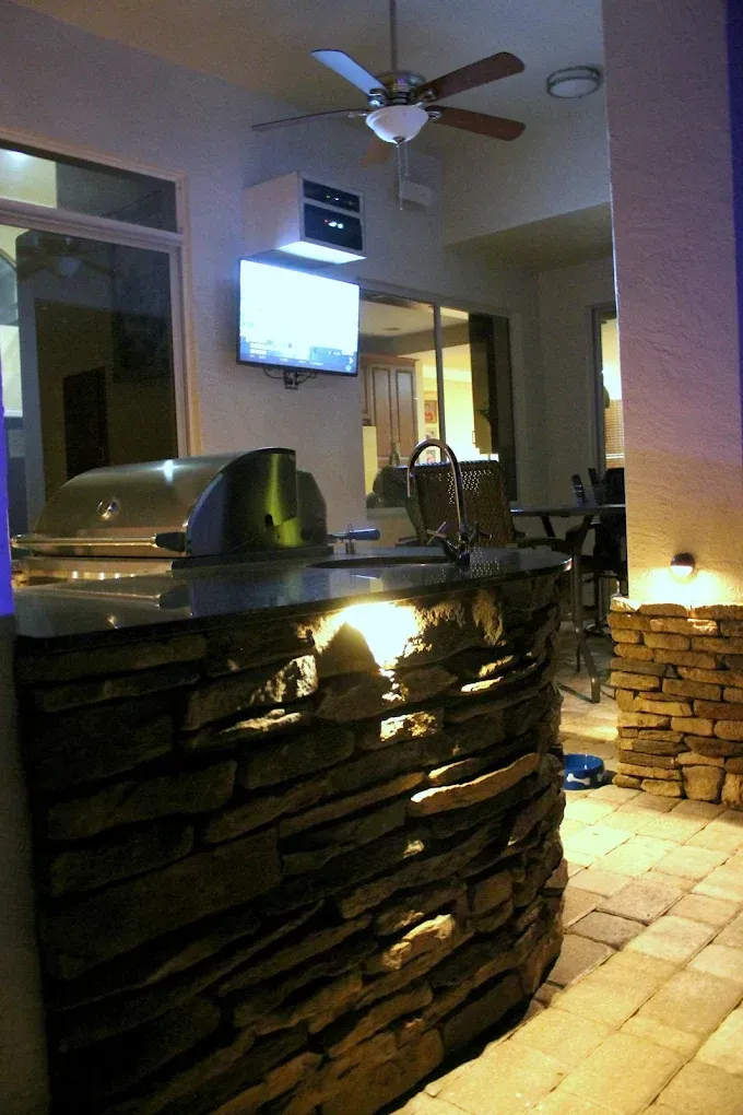 Outdoor kitchen with stone facade, grill, sink, mounted TV, and ceiling fan.