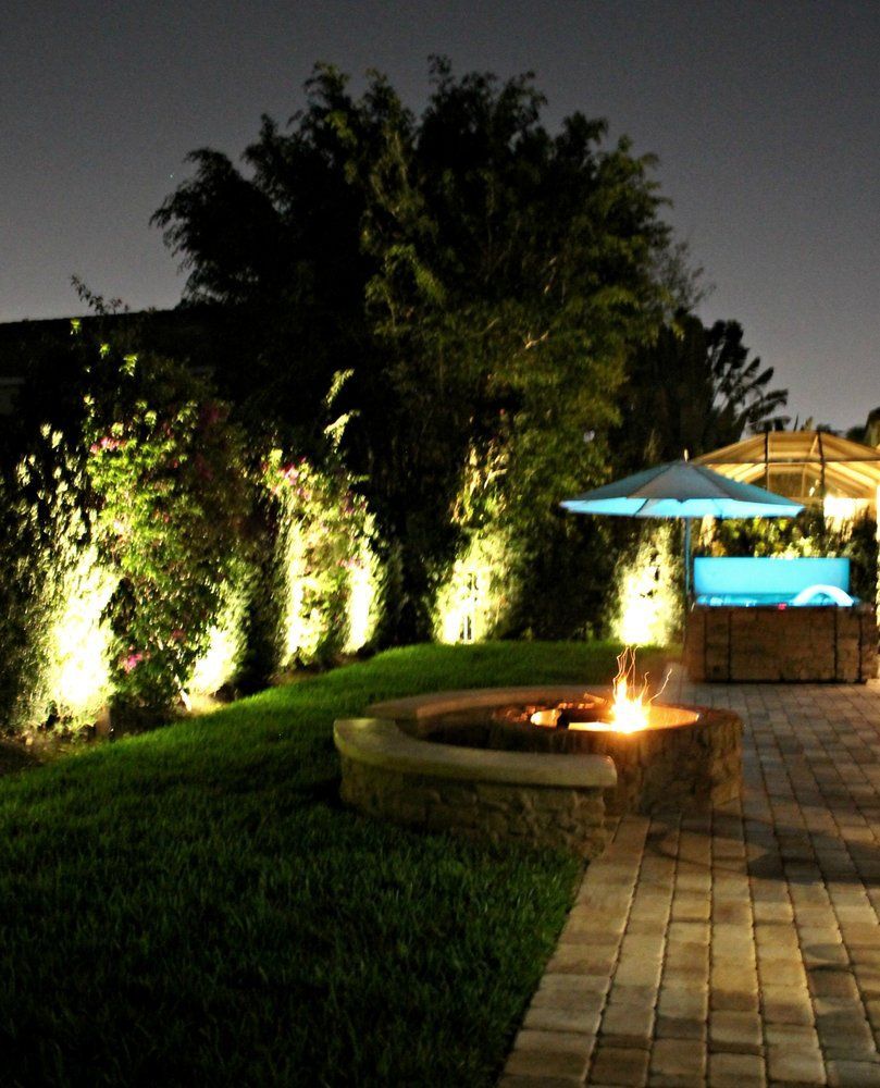 Nighttime backyard scene with lit fire pit, landscaping, and patio with umbrella.